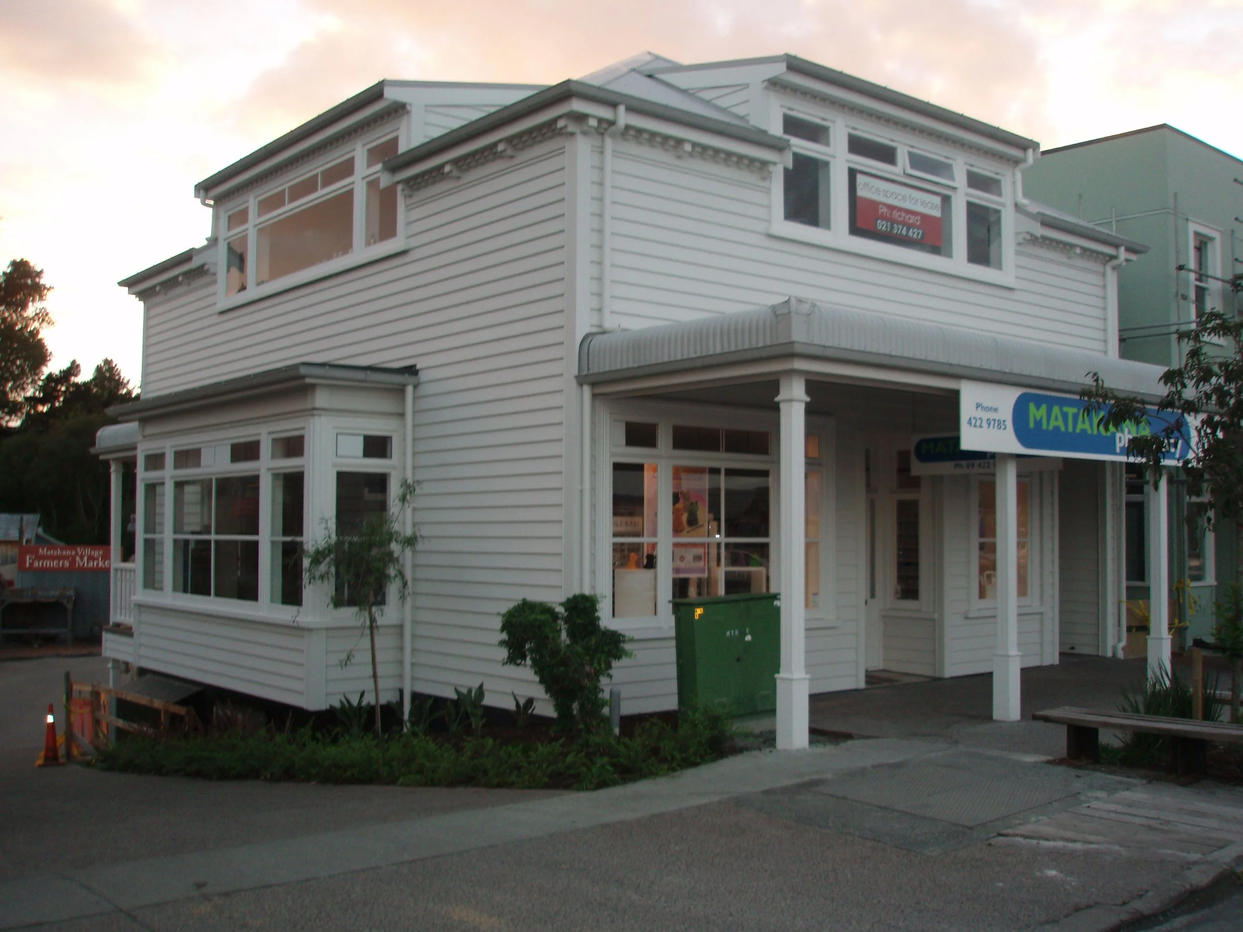 A two-story white building with large windows and a white awning over the entrance, with a sign indicating it is a business, possibly a real estate or office space, during sunset with a partly cloudy sky.