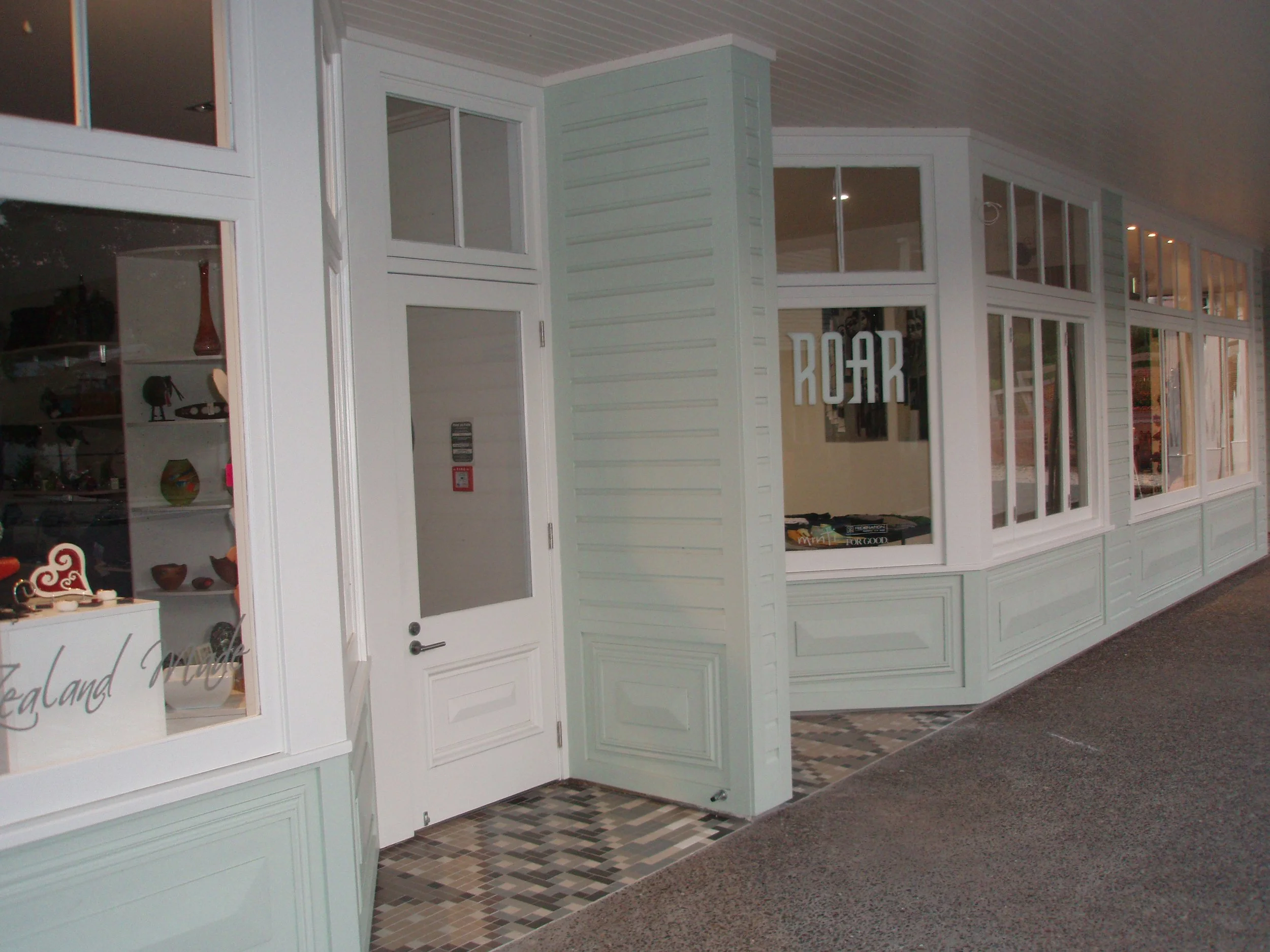 The image shows a storefront with large white-framed windows and a white door. The interior includes a display with various items, and the floor has a geometric tile pattern near the entrance.