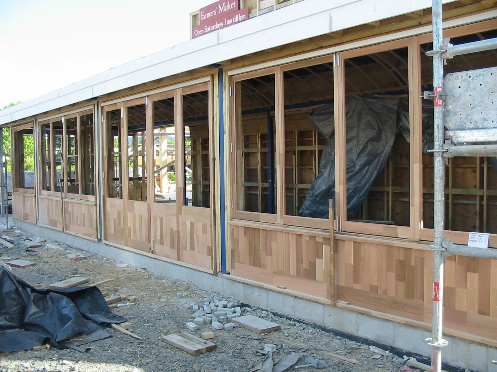 Wooden-framed building under construction with large windows and a sign indicating it is a farmers market open Saturday mornings. Construction materials and scaffolding in front.