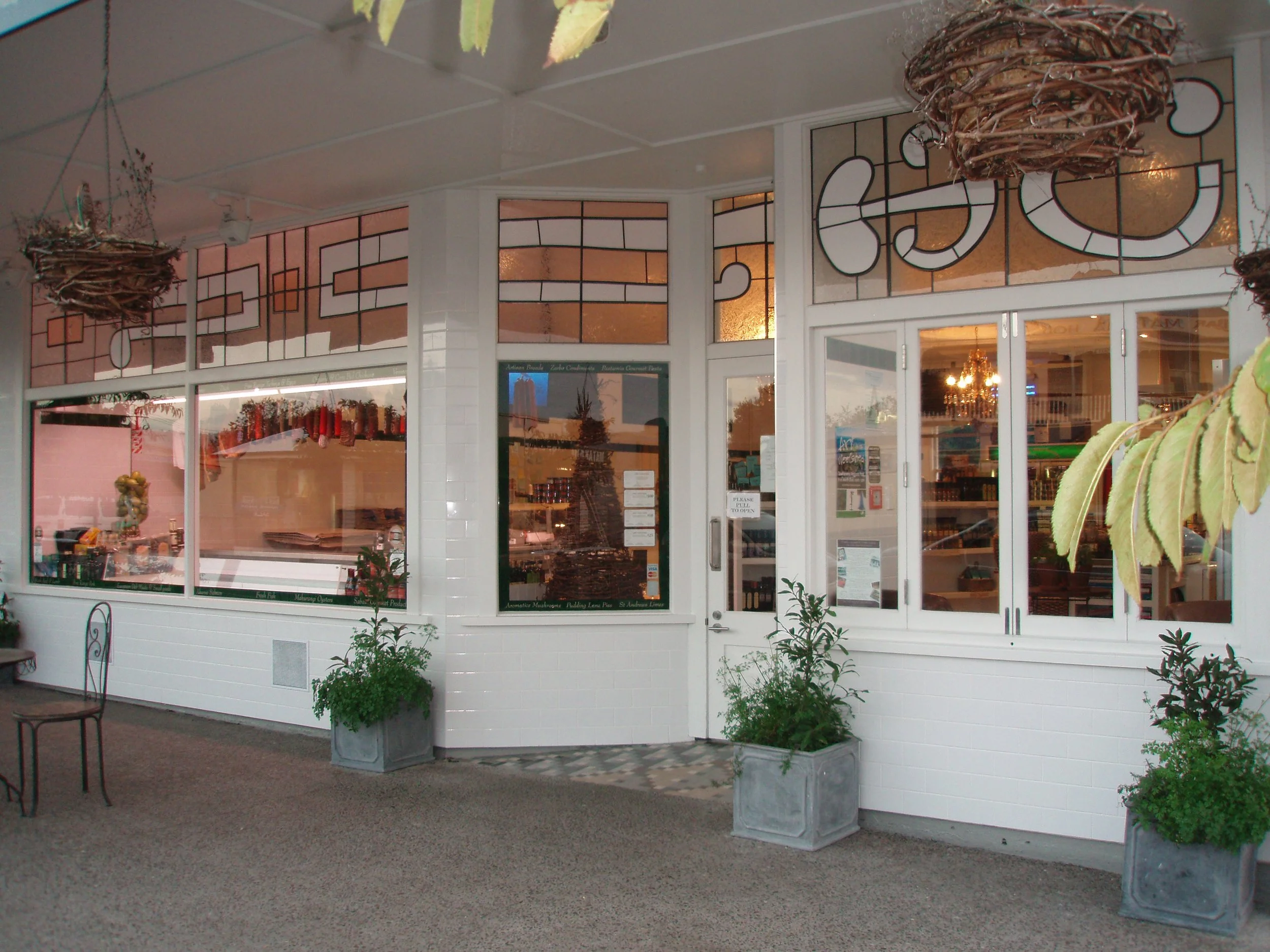 Exterior view of a storefront with large windows, potted plants outside, and stained glass panels in the upper part of the walls.