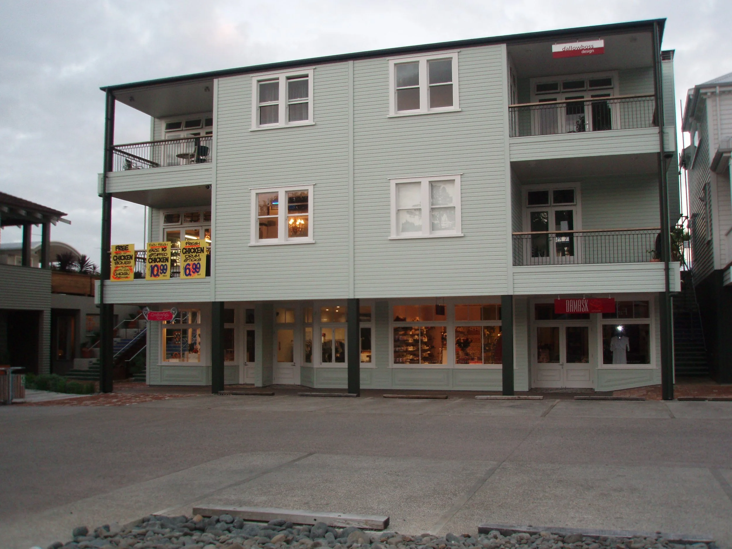 A three-story light green building with balconies and large windows, with advertisements for food on the ground floor, parked cars and a cloudy sky in the background.