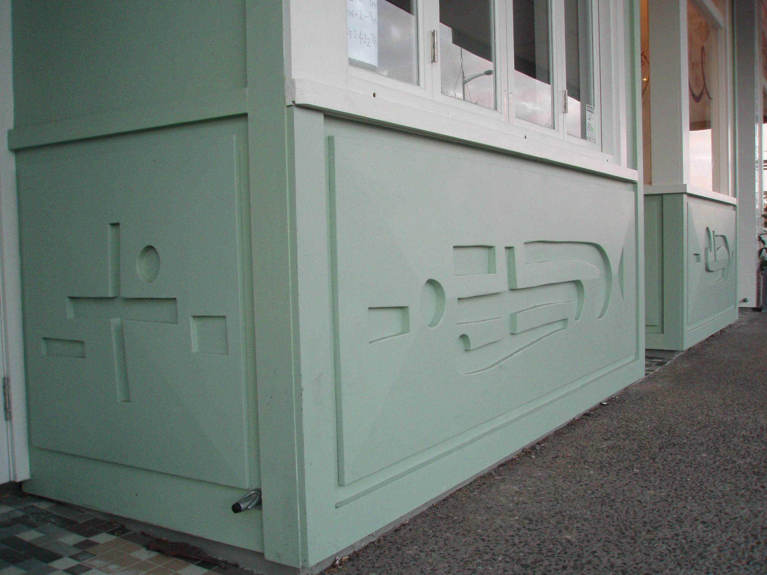 A close-up of a mint green painted building with decorative panels and a white-framed window. The building is situated on a concrete sidewalk, with a pattern of small tiles near the corner.