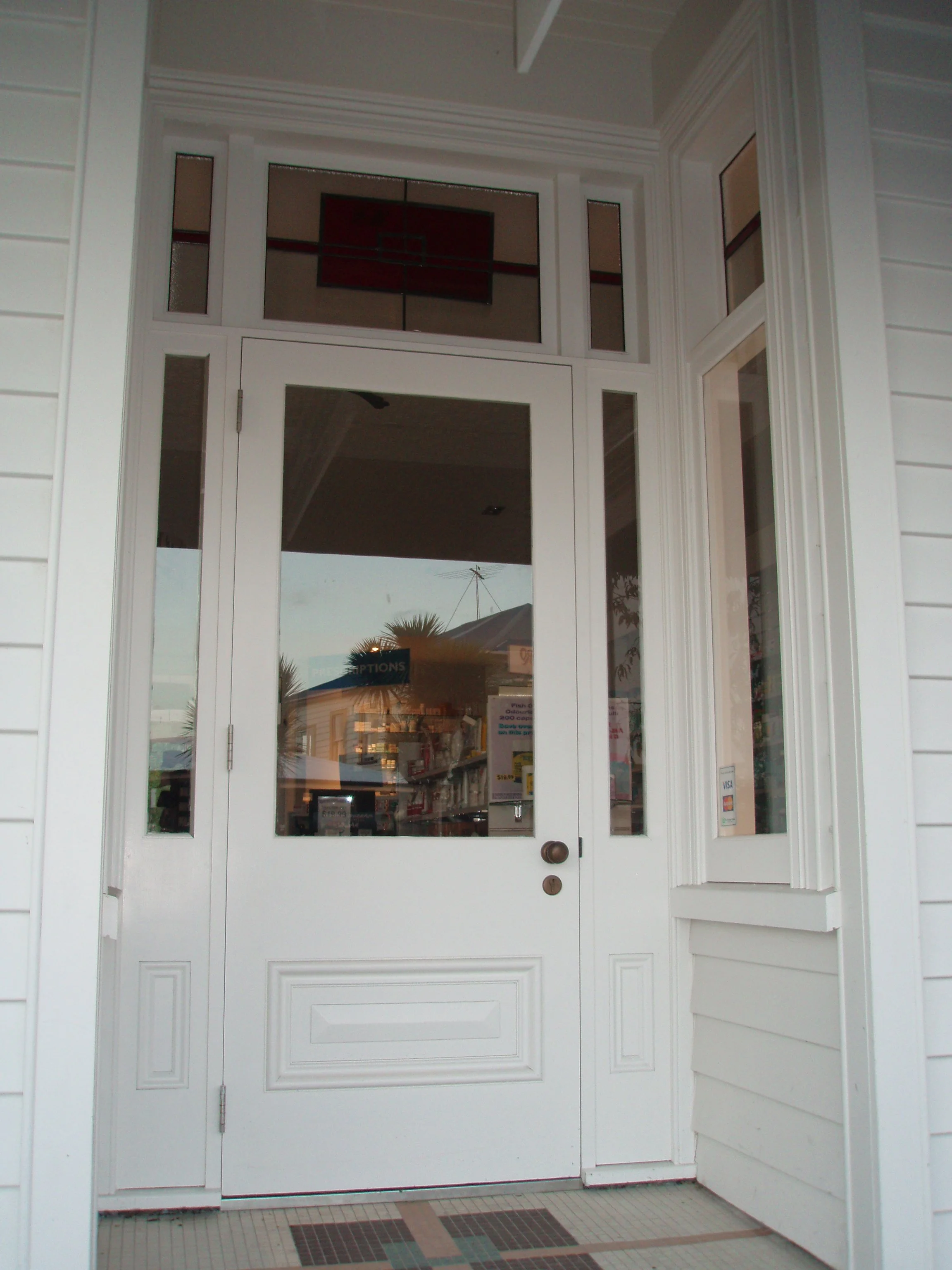 White front door with glass panels and a storm door, window on the right side, beige tile floor outside, and a partial view of a store interior through the glass.