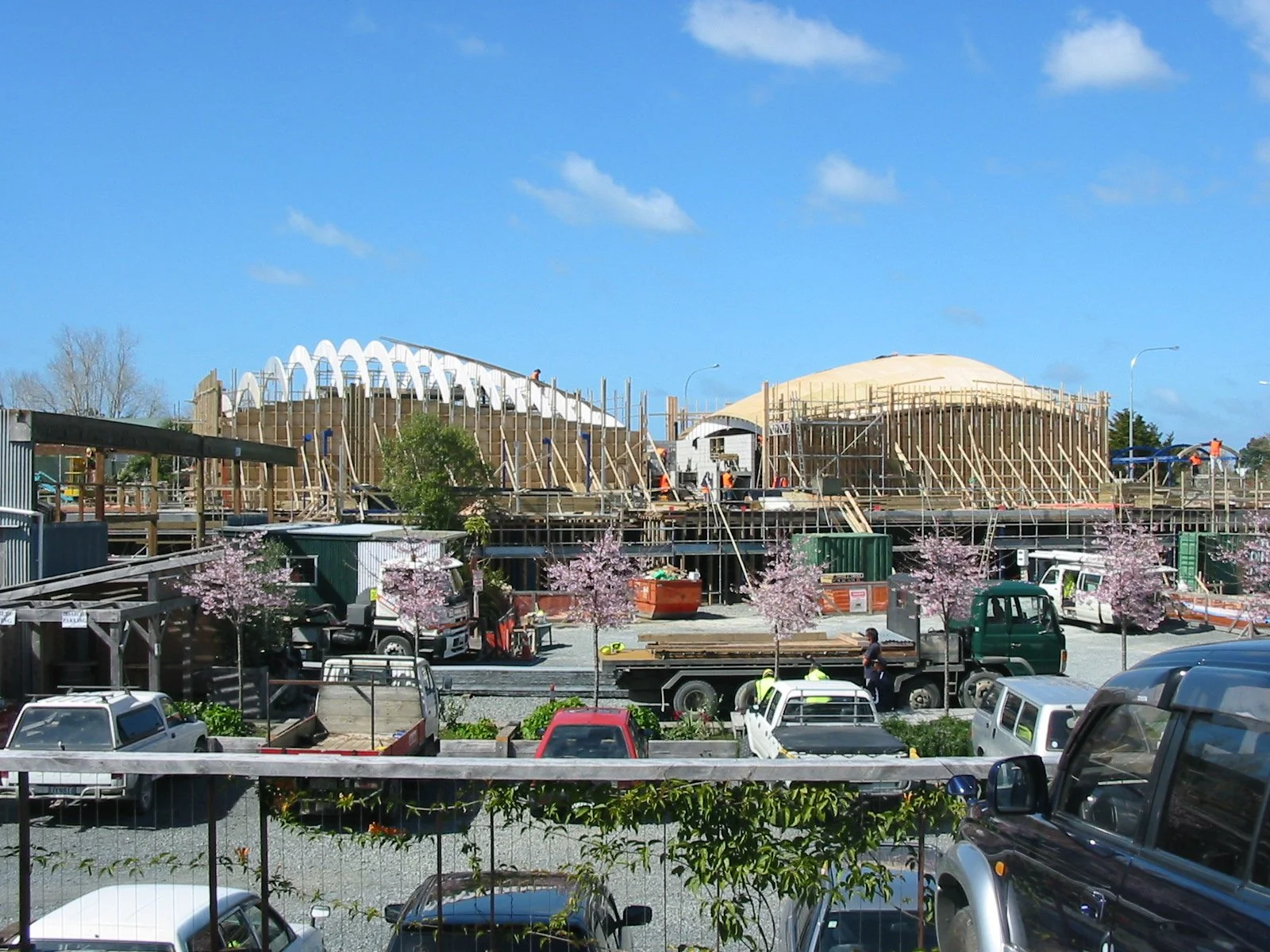 Construction site of a building with workers, partially built wooden structure, and a domed roof framework. There are trucks, cars, and pink flowering trees in the foreground, under a clear blue sky.