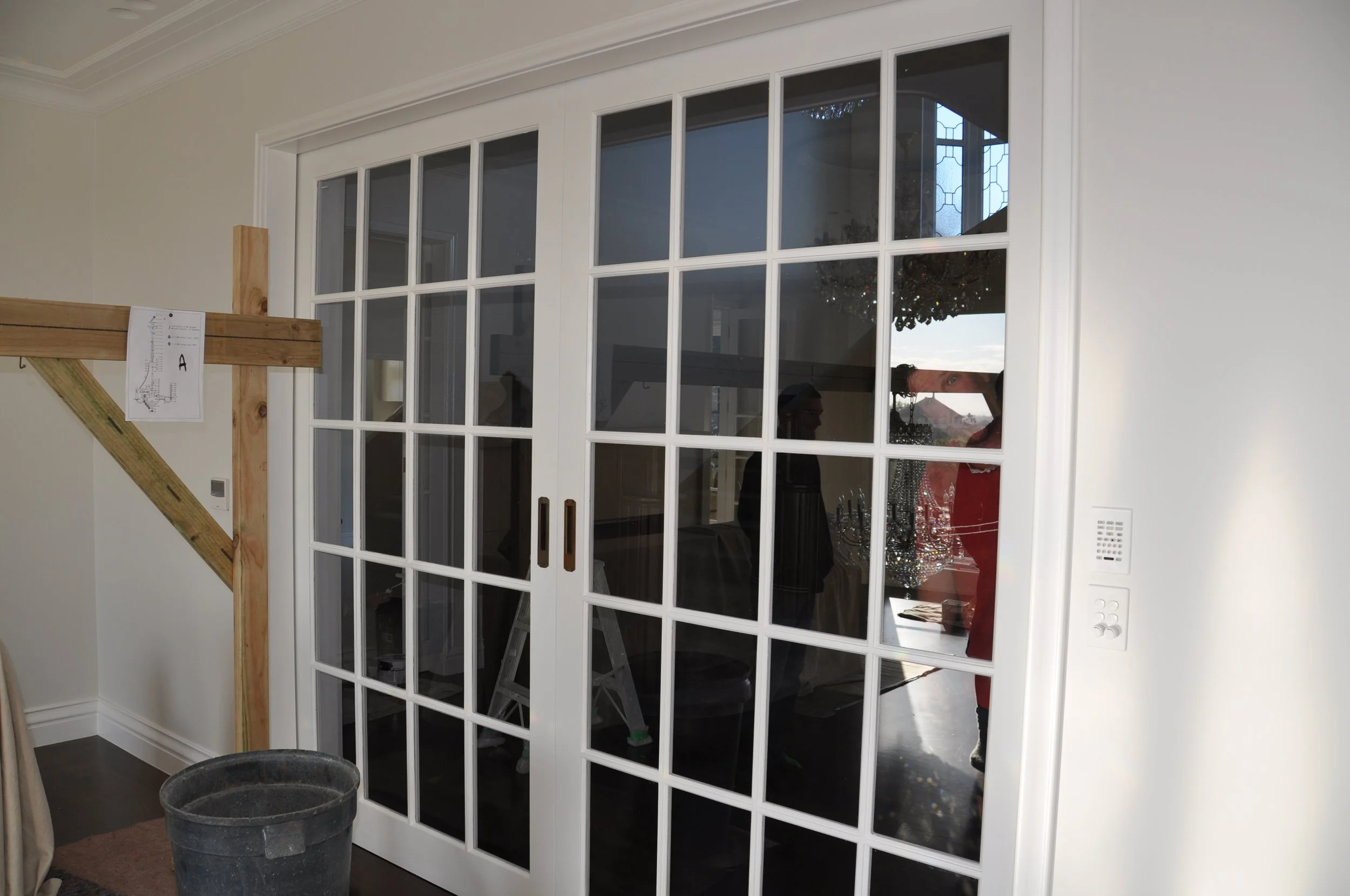 White sliding glass door with multiple square panes, leading to an interior room with Christmas decorations and a chandelier, in a home during daytime.