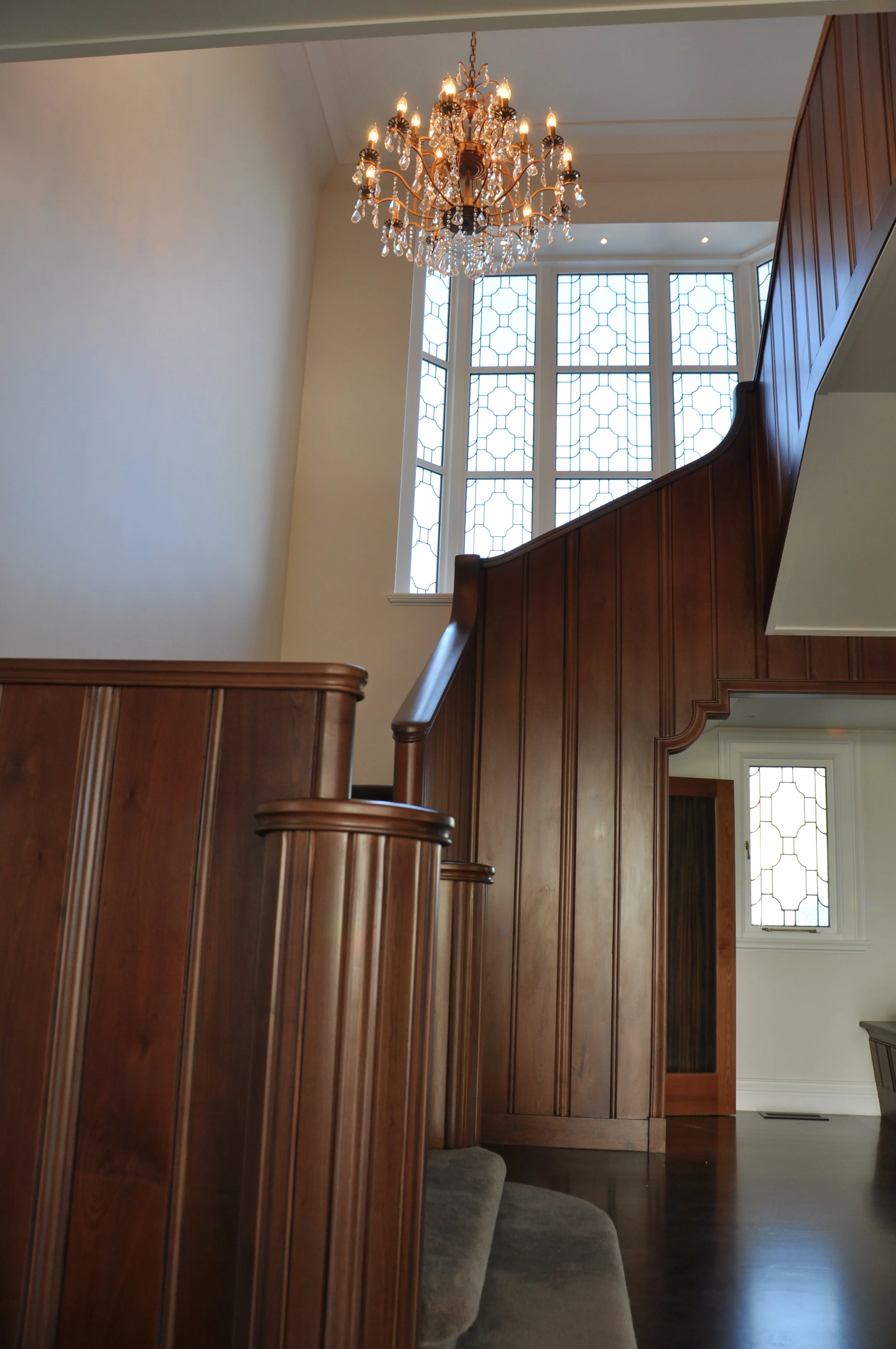 Interior view of a staircase with wood paneling and a large window with geometric patterns, a chandelier hanging from the ceiling, and a partial view of a door with a similar window pattern.