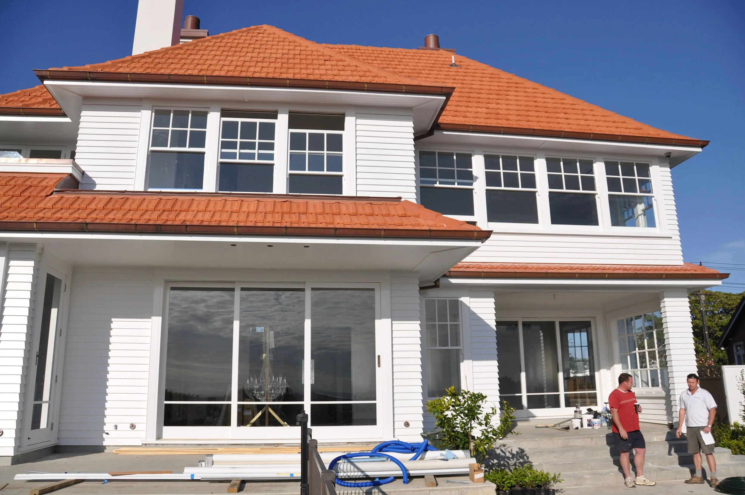 A large two-story house with a red tiled roof and white siding. The house has large windows, and there are two men standing on the patio in front of it, one in a red shirt and dark shorts, and the other in a white shirt and khaki pants. Construction 