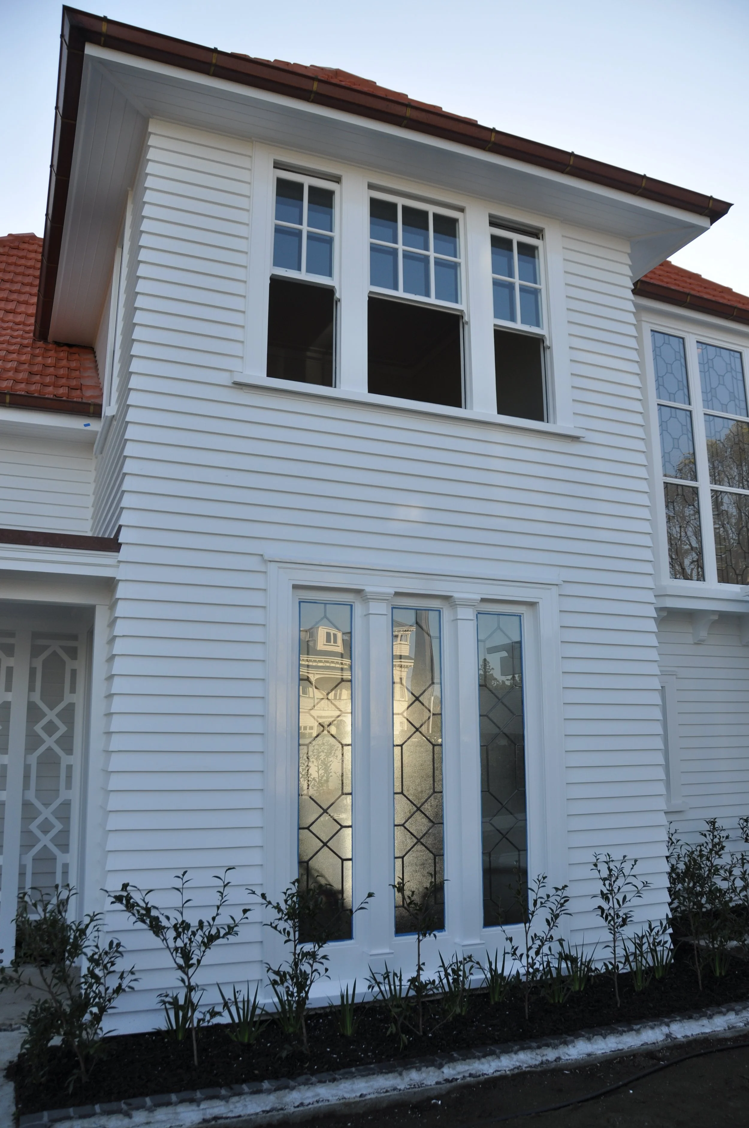 White house with a large window on the upper floor, decorative glass door on the lower floor, white siding, and a flower bed in front.