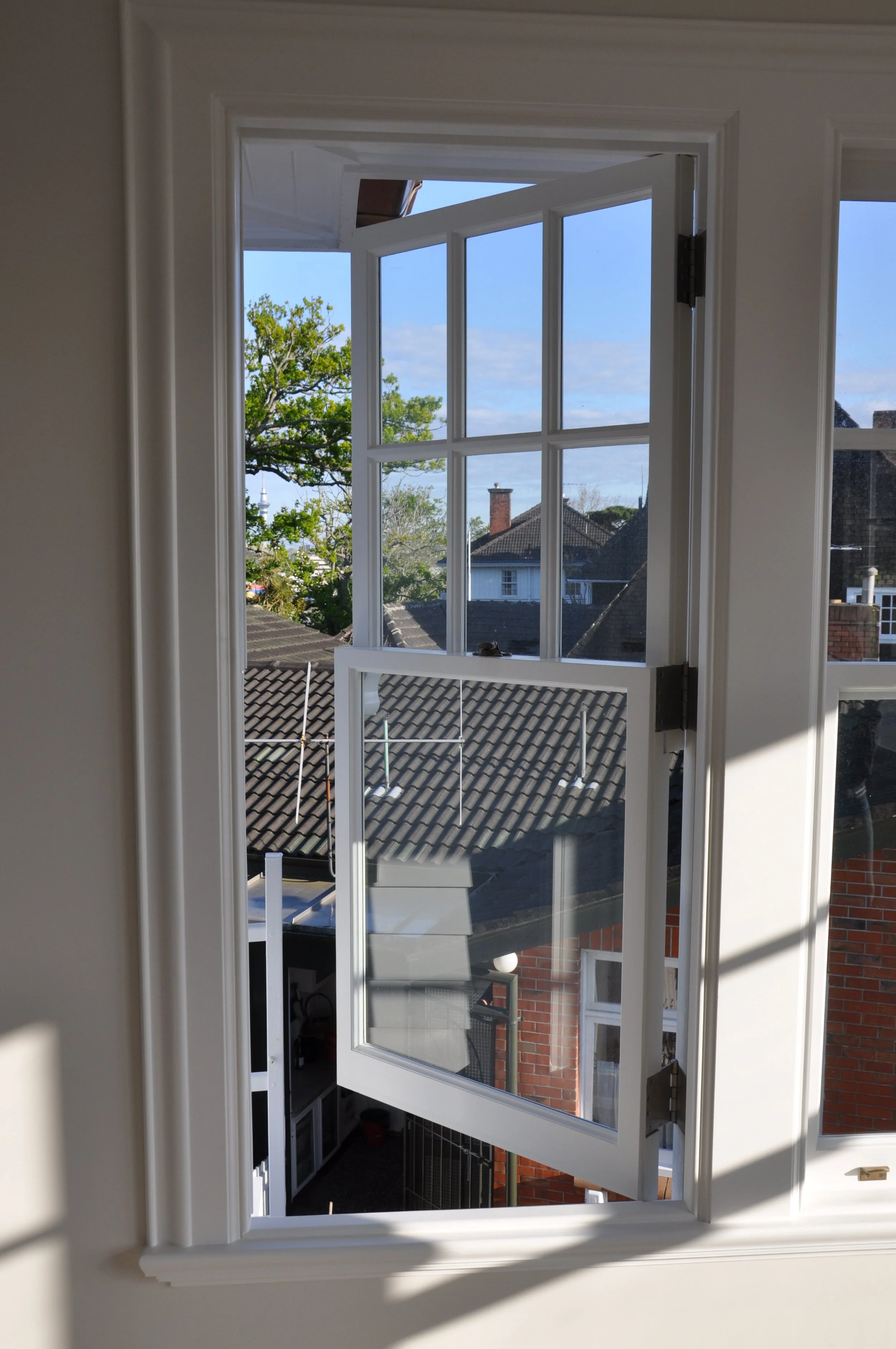 Open white window with multiple panes, view of rooftops, trees, and blue sky with some clouds outside.