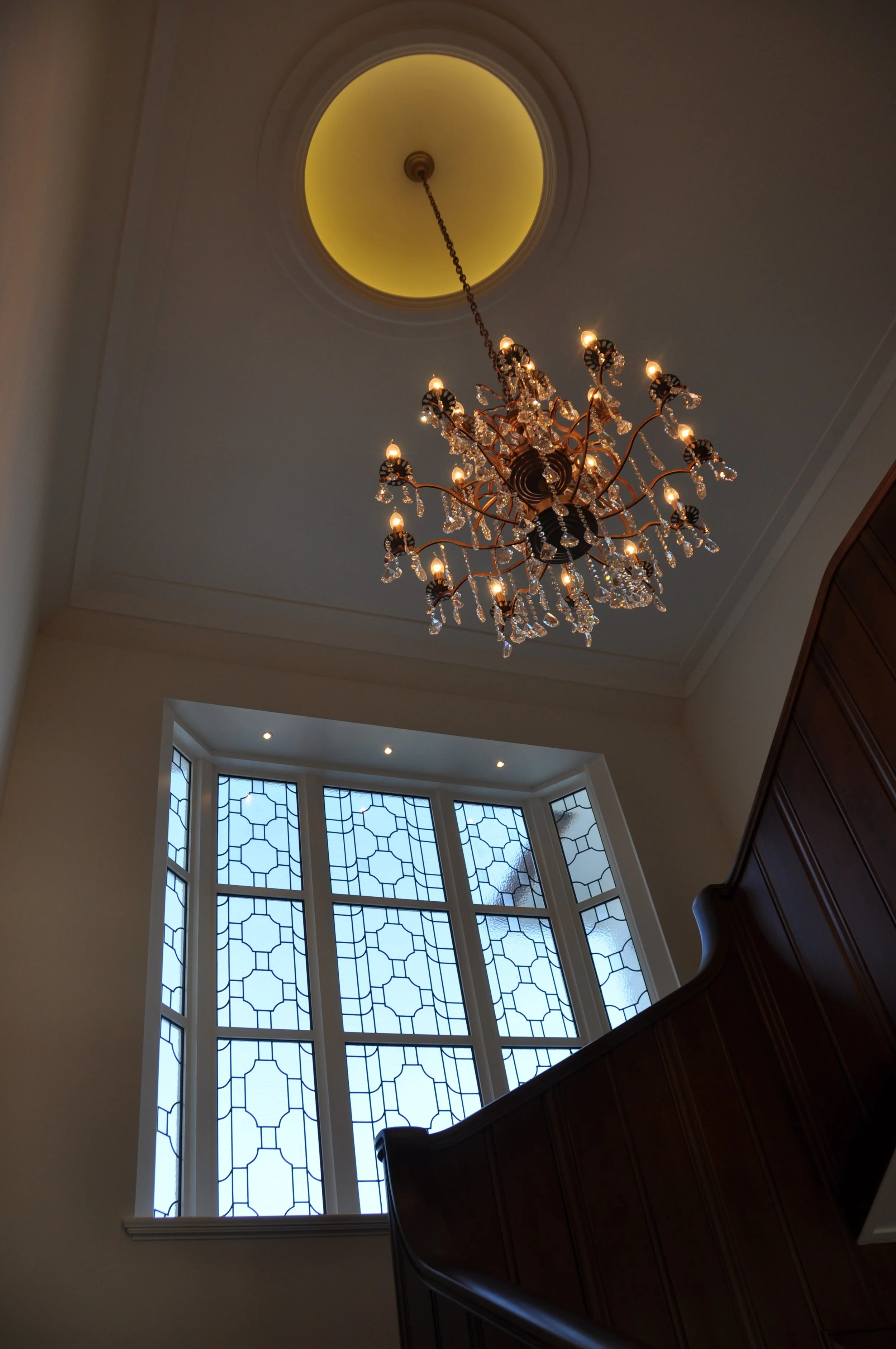 Interior of a room with a chandelier hanging from the ceiling, a large window with geometric patterned panes, and a wooden staircase railing.