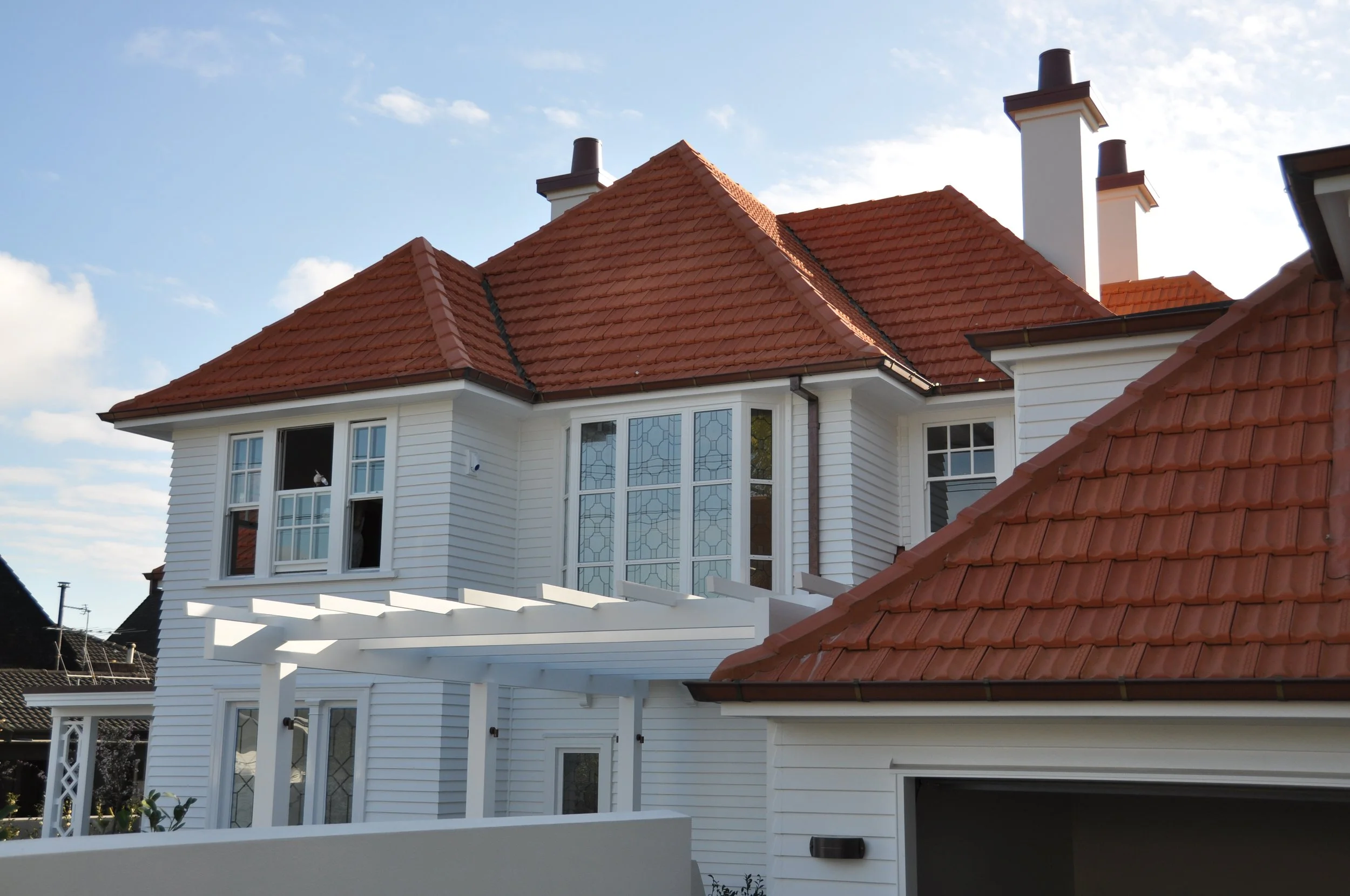 A multi-story house with white siding and a red tile roof, featuring large windows with decorative panes and a covered patio area underneath.