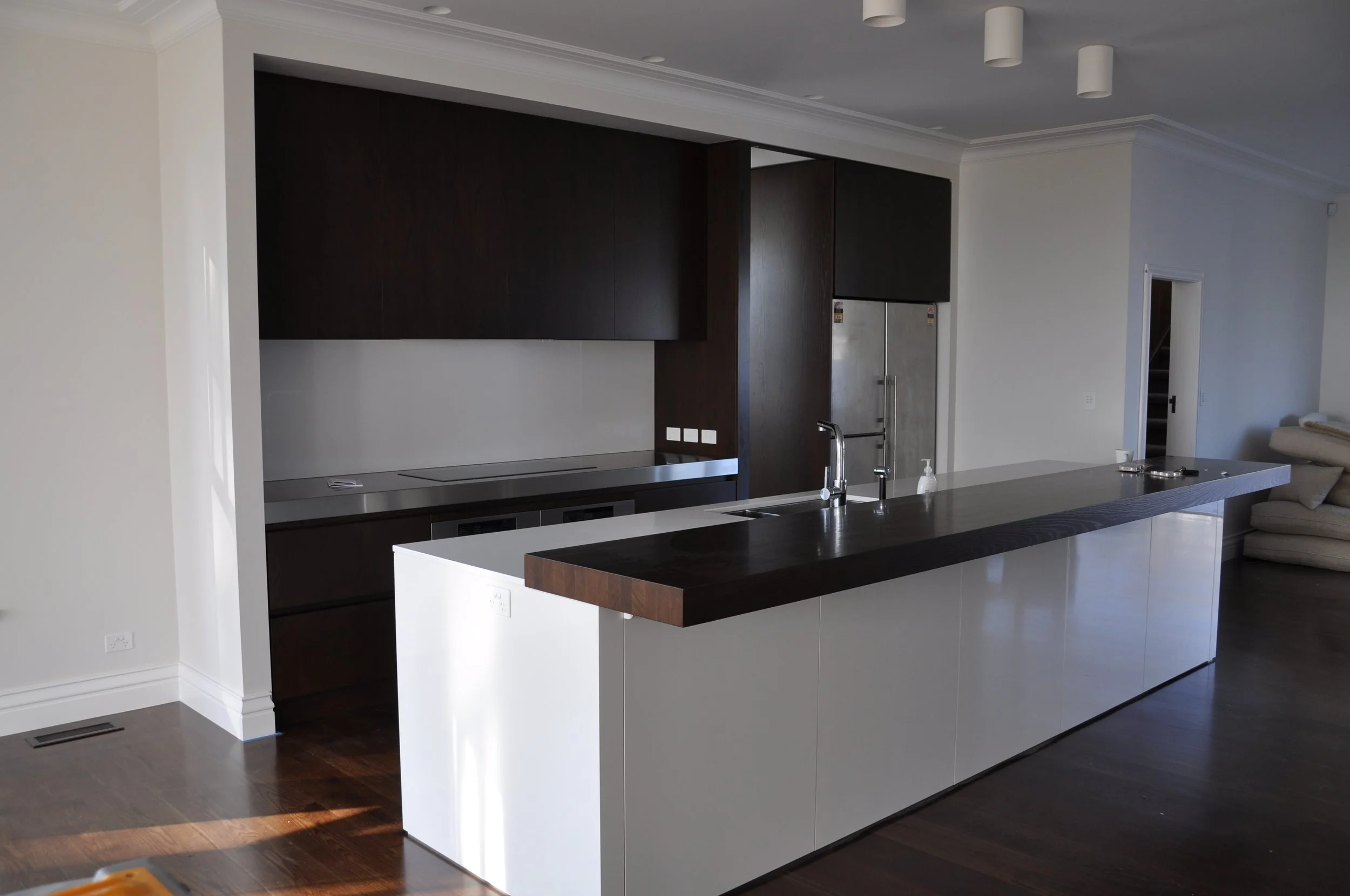 Modern kitchen with a white island, dark wood cabinets, stainless steel refrigerator, and a living area with a beige sofa.