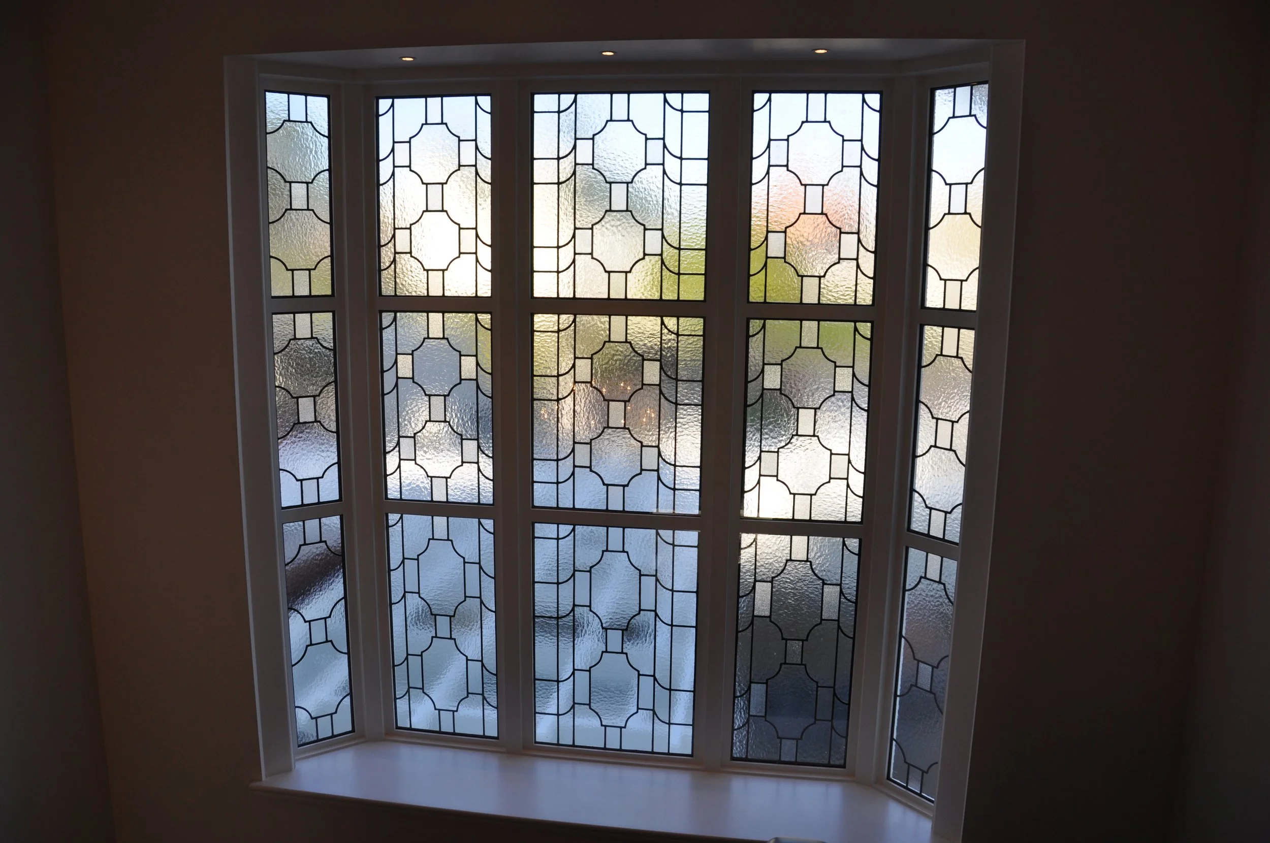 A bay window with a geometric patterned stained glass design, looking outside at a slightly cloudy sky and trees.