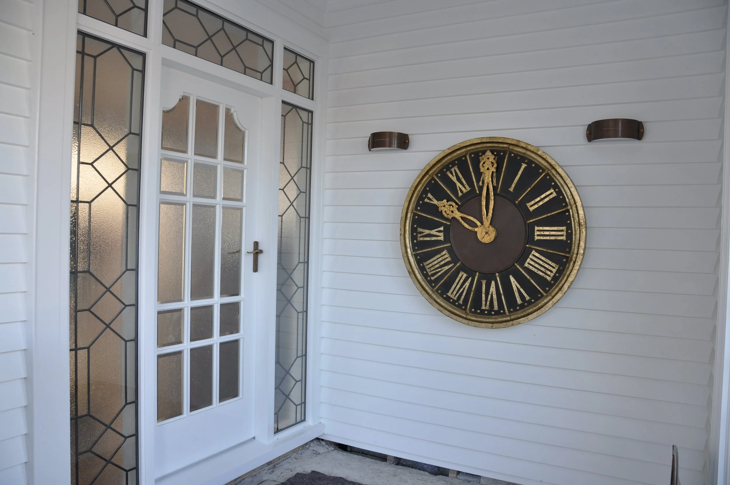 Front porch with white siding, glass front door with geometric stained glass, and a large round black and gold clock on the wall.