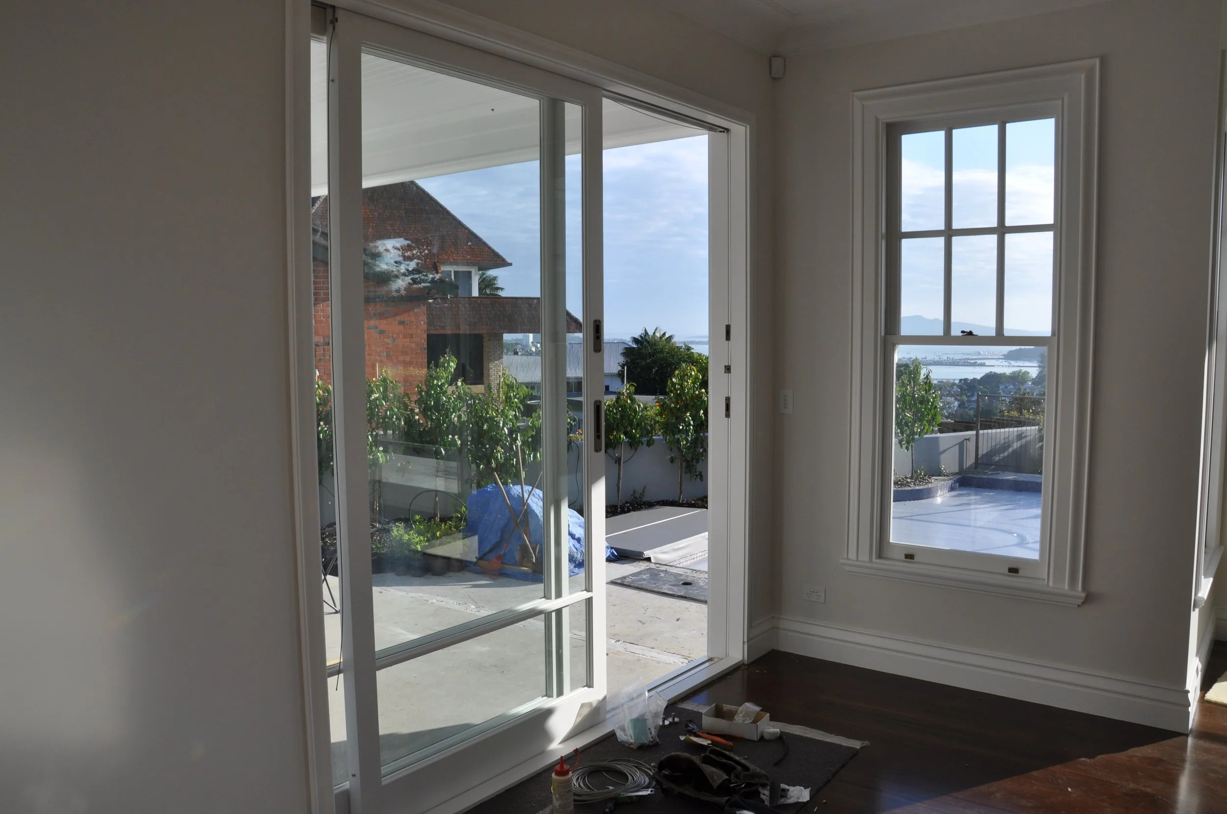 Interior room with sliding glass door leading to an outdoor balcony with plants, and a large window showing neighboring houses and a water body in the distance.