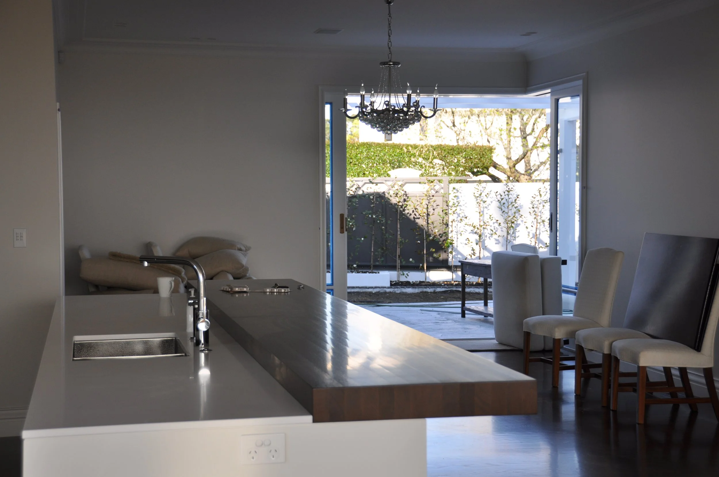 Inside view of a modern kitchen with an open sliding glass door leading to a backyard with green bushes and trees, dining table, chairs, chandelier, and a countertop with a sink.