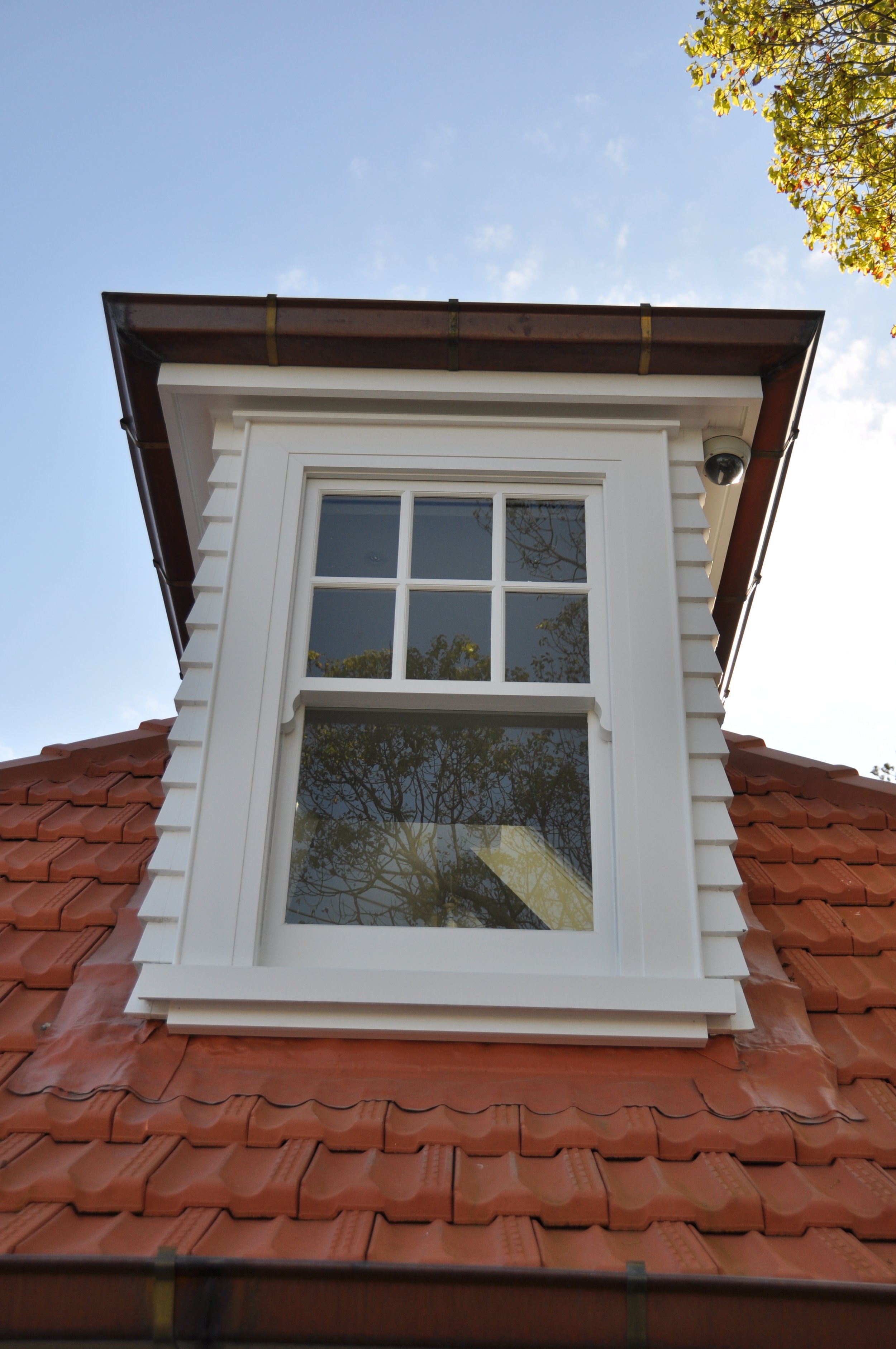 Close-up of a dormer window with a white frame on a red tiled roof, with a smoke detector located near the top right corner of the window. The sky is clear and blue, with trees reflected in the window.