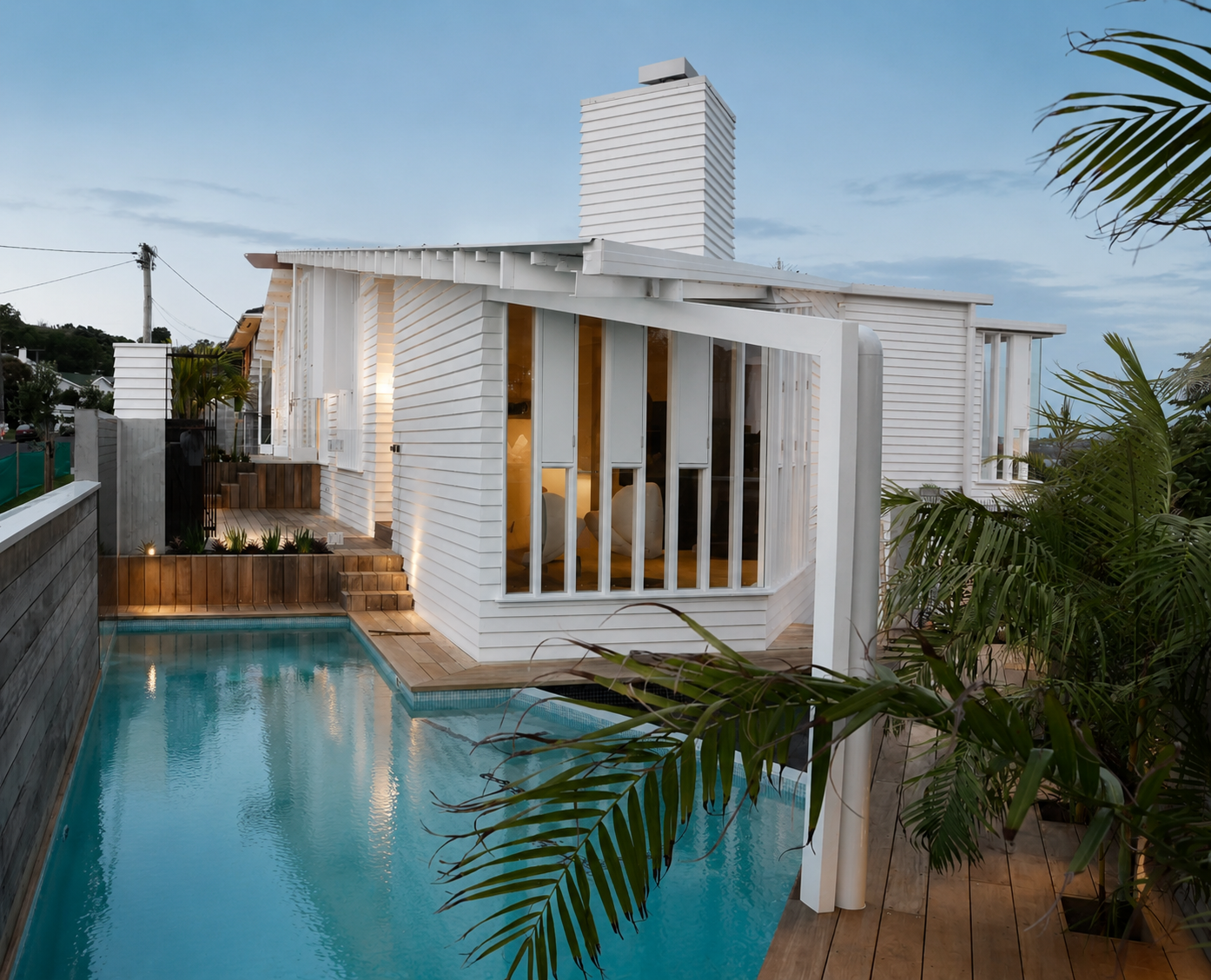 Modern white house with large windows, surrounded by a swimming pool and lush green plants, during dusk.