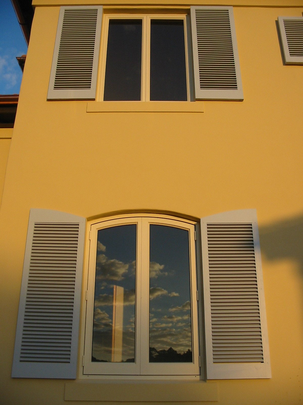 A yellow building with white-framed glass doors and windows, featuring white shutters, reflecting a cloudy sky.