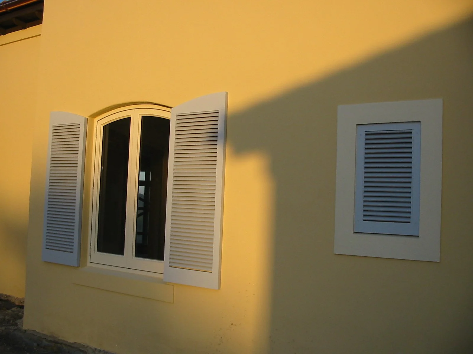 Yellow exterior wall of a house with a large arched window and white window shutters, illuminated by sunlight.