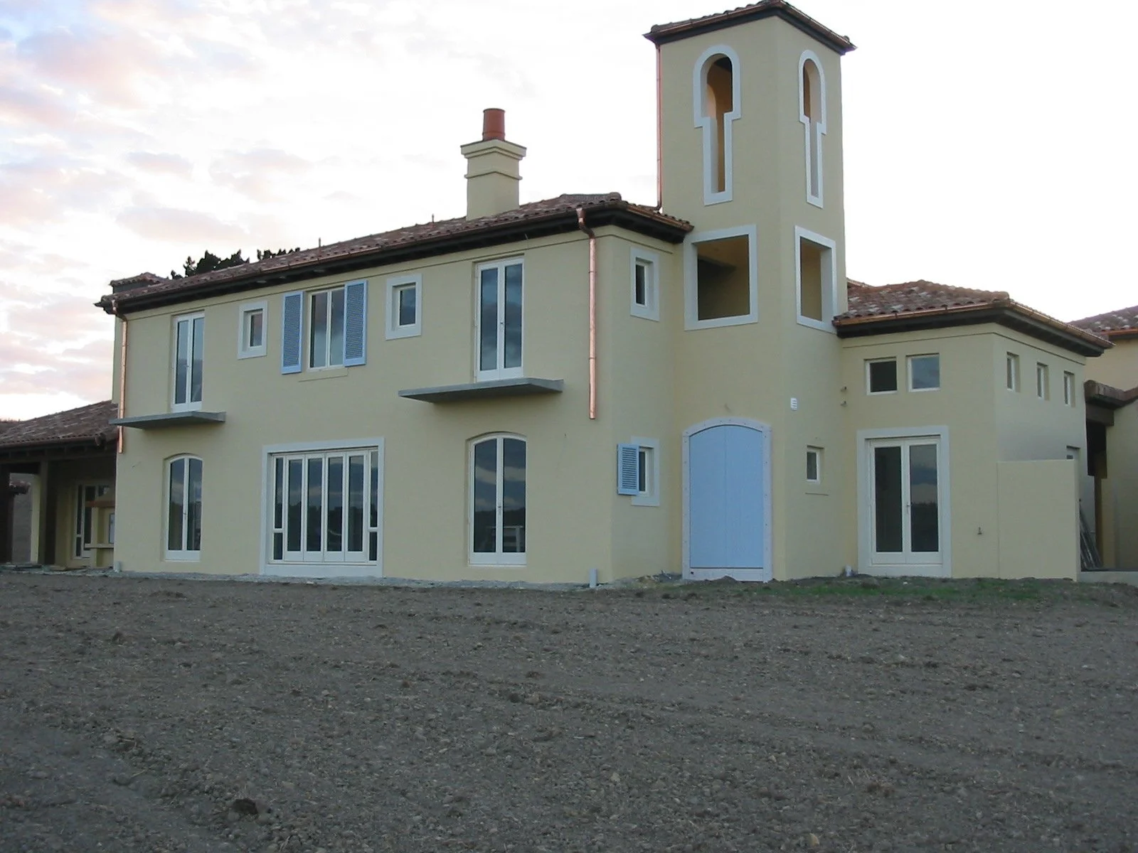A yellow house with multiple windows and a tower on the right side, under a cloudy sky, with a dirt ground in the foreground.
