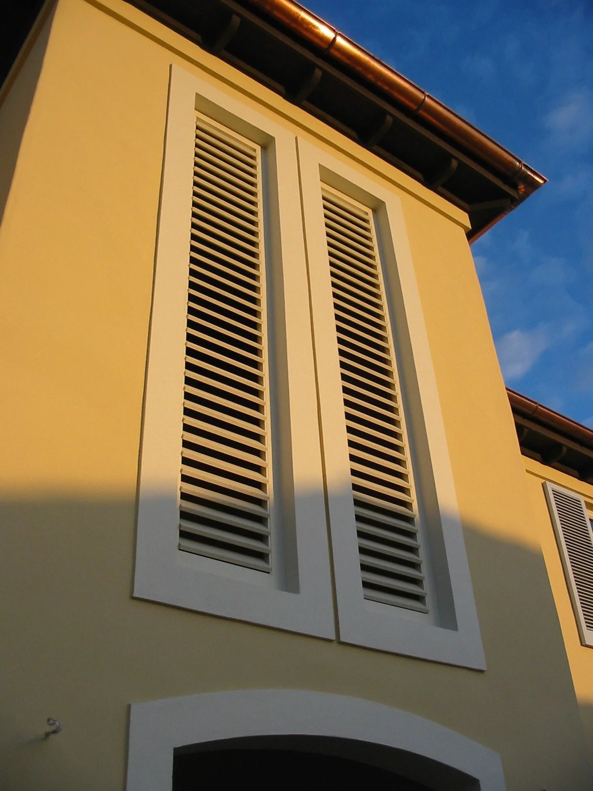 Close-up of a yellow building facade with white-edged louvered shutters and a curved archway at the bottom, under a clear blue sky.