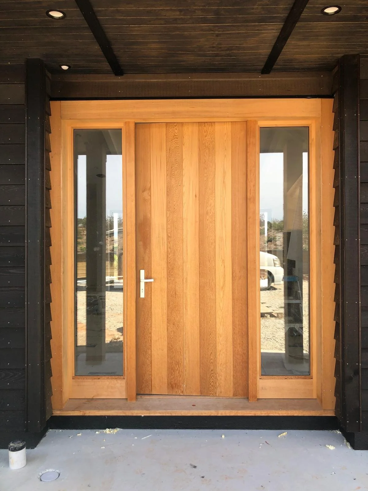 Front view of a wooden door with glass panels on each side, installed in a house with black siding and a wood ceiling.
