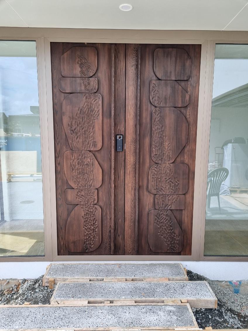 Wooden front door with carved abstract patterns, flanked by glass panels, with a small step and unfinished ground in front.