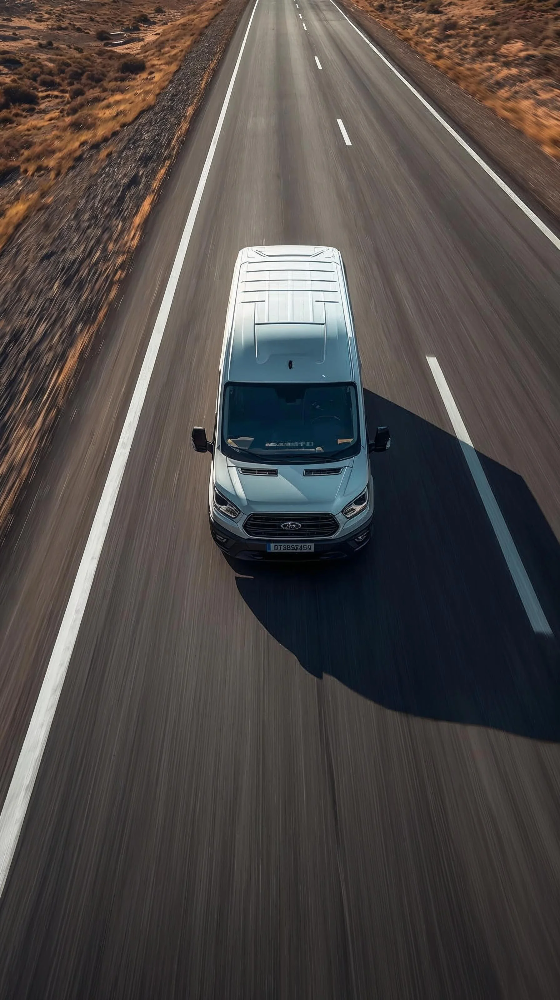 A silver Ford van driving on a two-lane highway through a desert landscape with brown and orange vegetation.