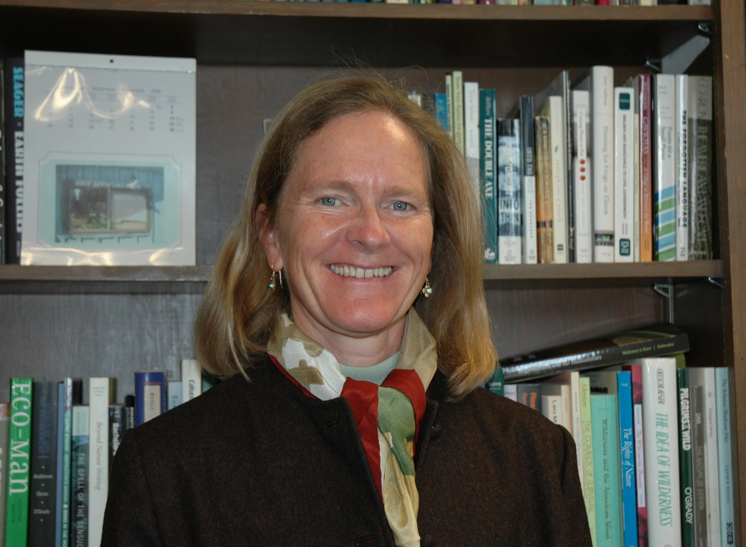 Rae Raptor with blonde hair smiling, standing in front of a bookshelf filled with various books.