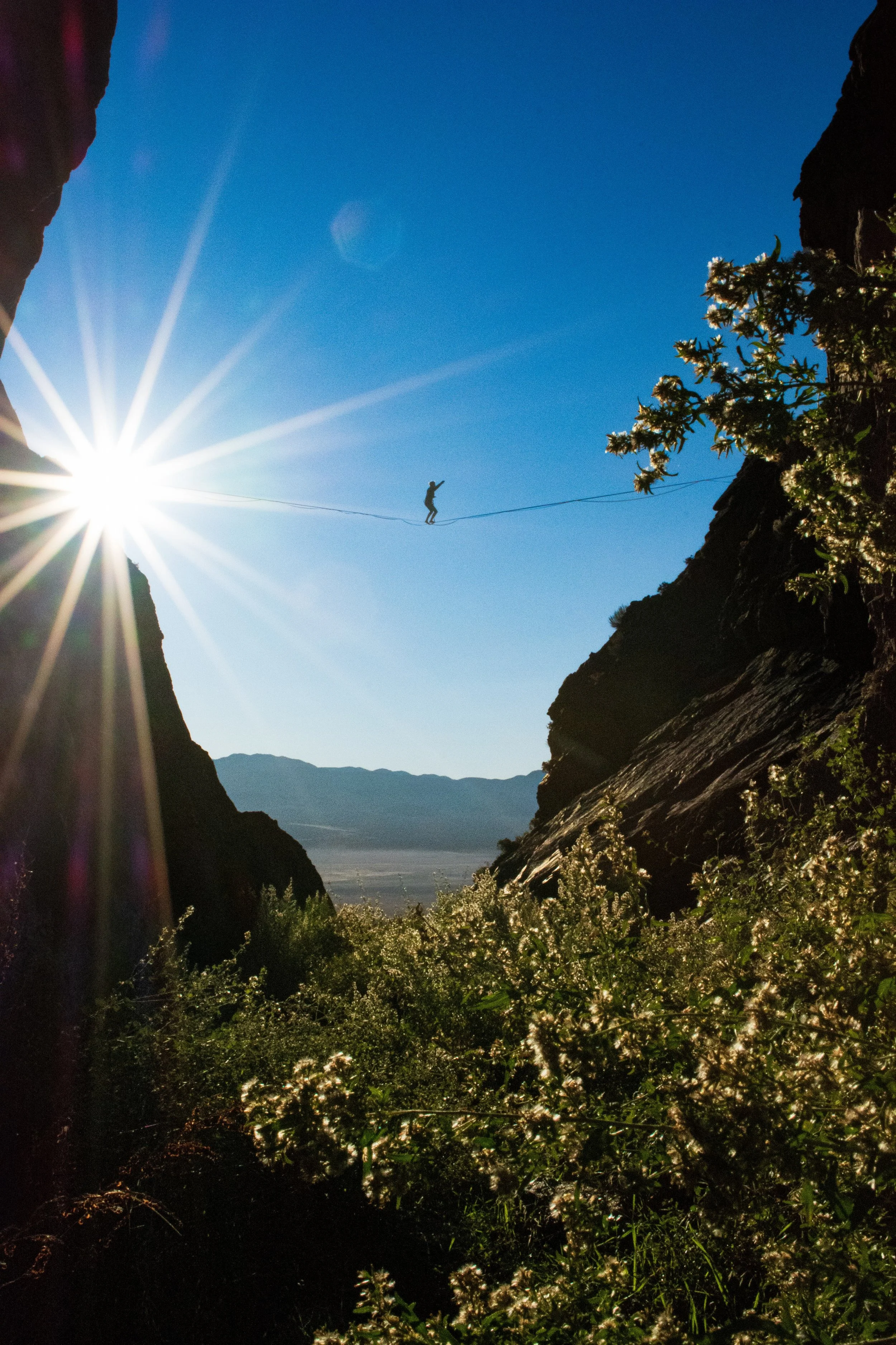Rae Raptor  walking on a highline between two rocky cliffs during daytime, with the sun shining brightly in the sky and creating lens flare.