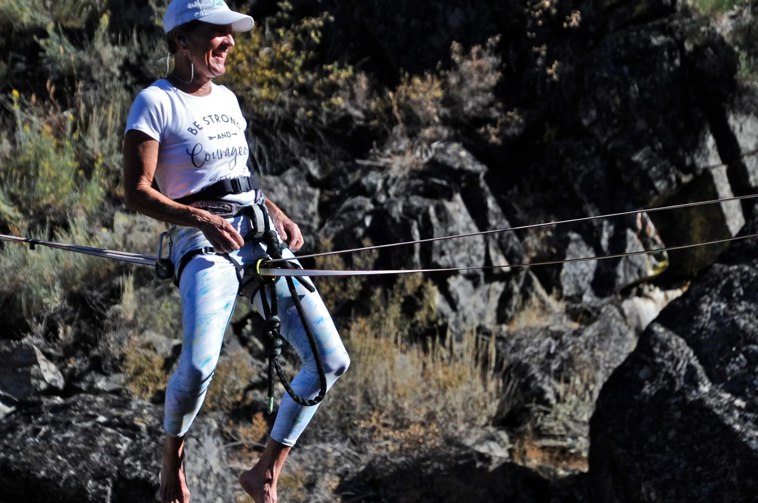 Rae Raptor in a white baseball cap, white tie-dye leggings, and a white T-shirt with the words 'Be Strong and Courageous' walks on a narrow mountain trail on a suspension bridge with cable rails, surrounded by rocky terrain and sparse vegetation.