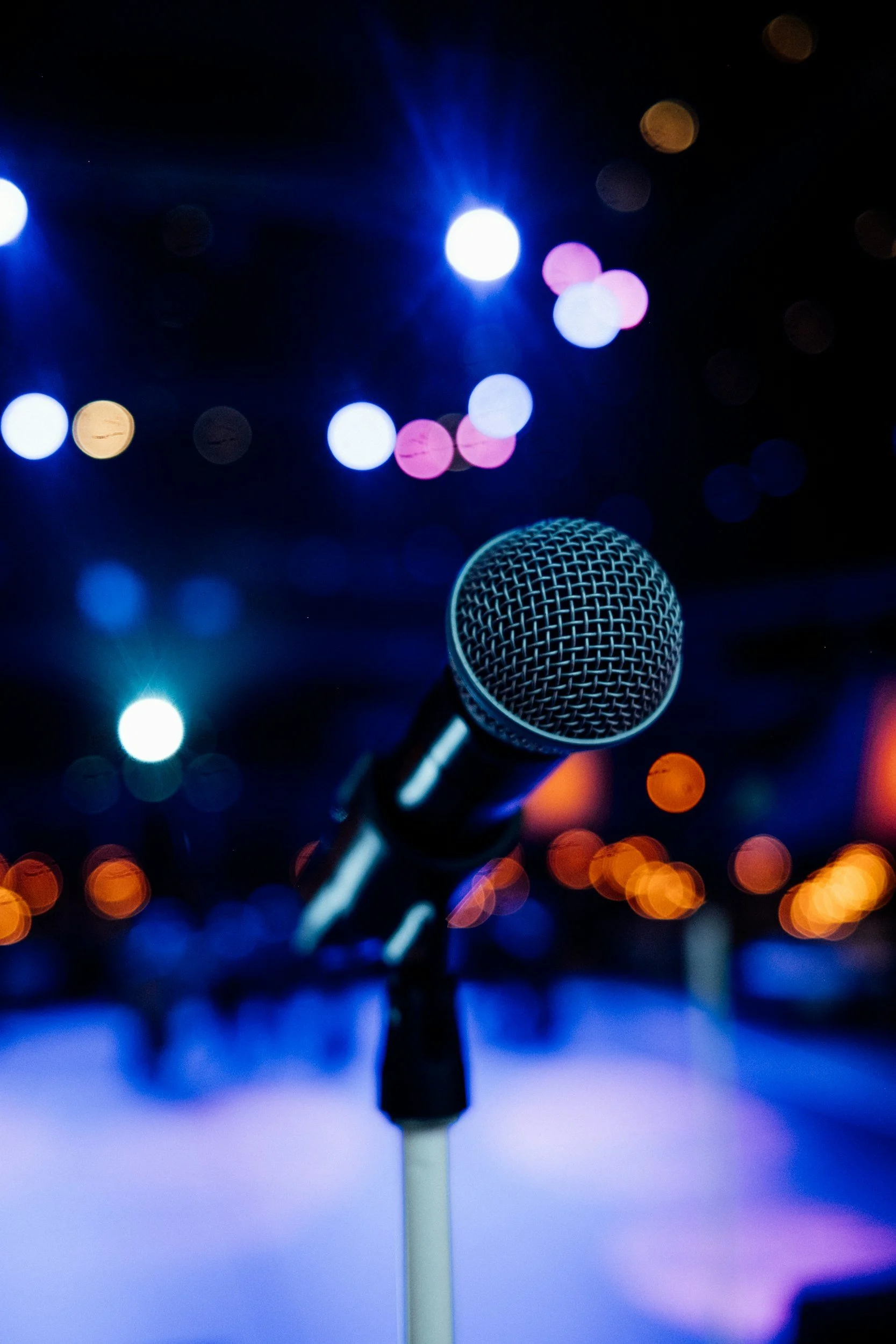 Close-up of a microphone in front of colorful blurred stage lights, suggesting a performance or speaking event.