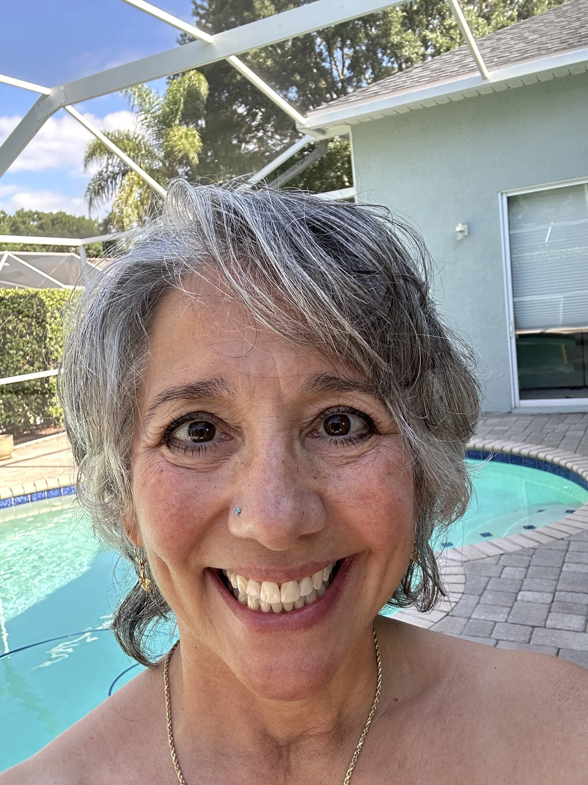 A woman with short, gray hair and a nose piercing smiling outdoors near a swimming pool, with a house and green trees in the background.