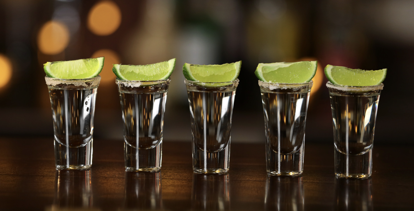 Five tequila shots in shot glasses garnished with lime wedges, lined up on a wooden surface.