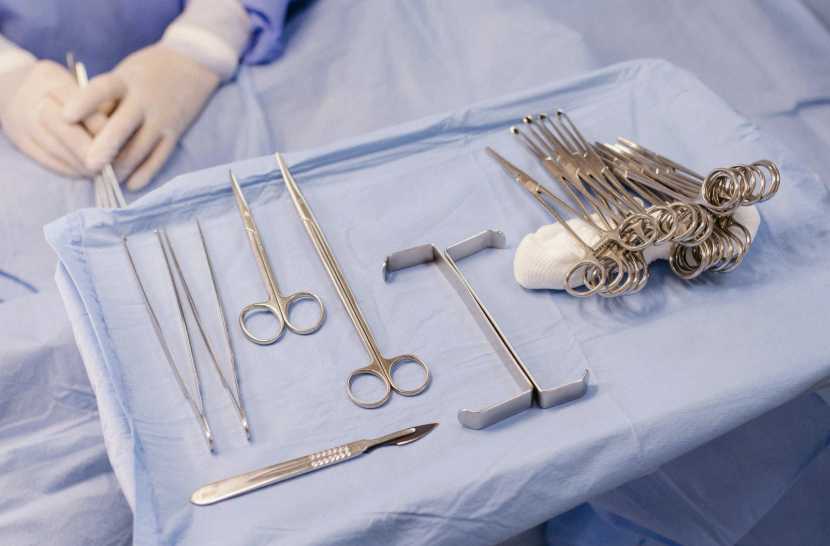 Medical surgical instruments, including forceps, scissors, and clamps, are arranged on a blue sterile drape in a surgical setting.