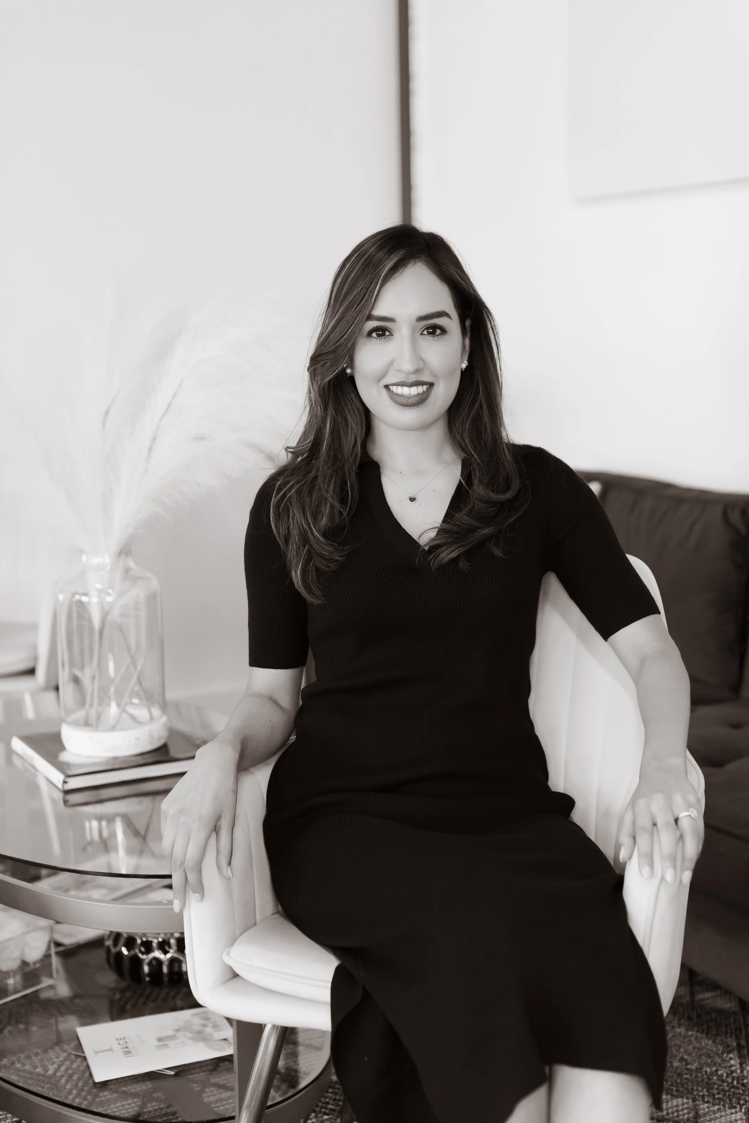 A woman with long dark hair, wearing a black dress, sitting on a white armchair, smiling, in a modern indoor setting with a side table, books, and a decorative vase with pampas grass in the background.