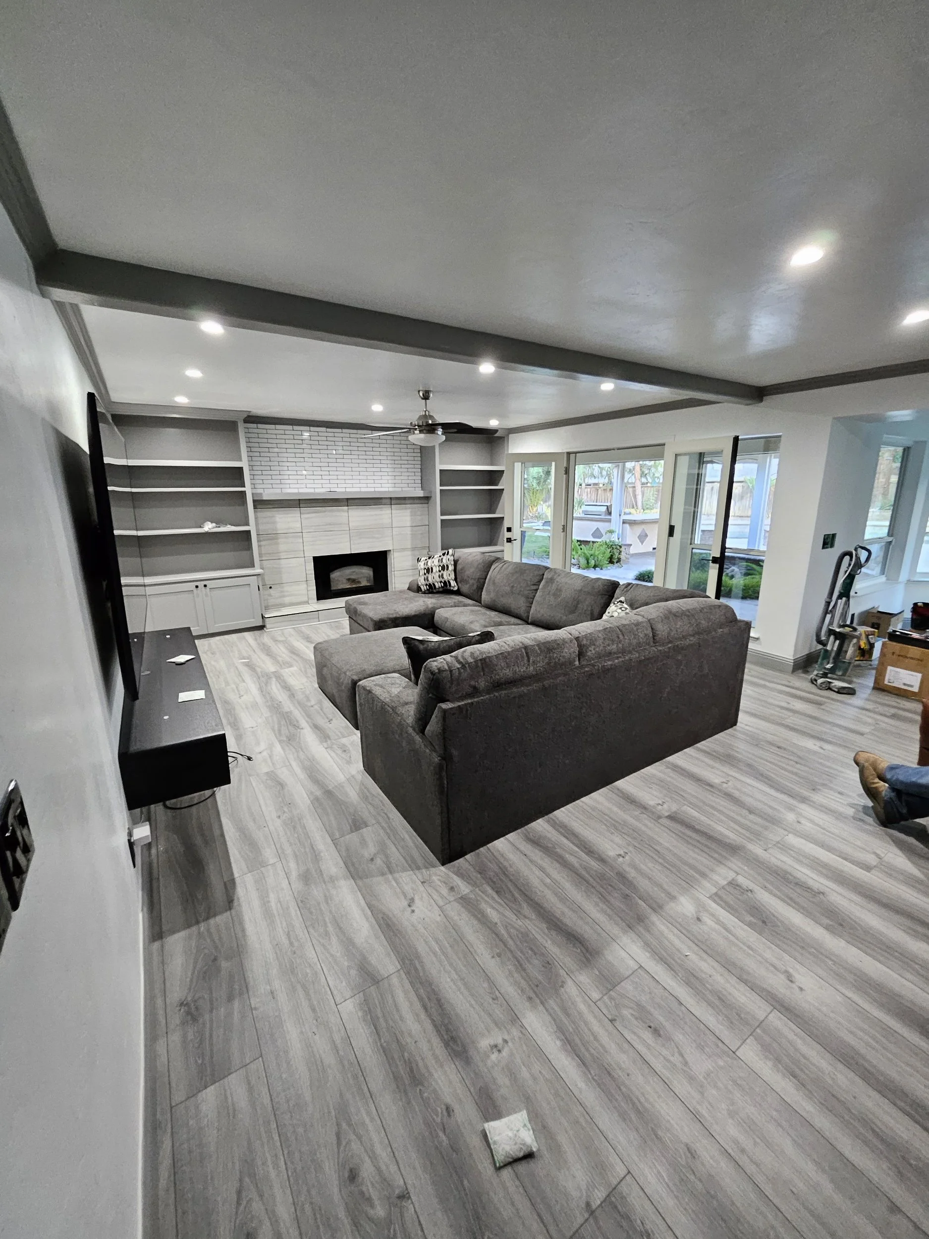 Living room with gray sectional sofa, built-in shelves, a fireplace, large windows with views of a backyard, ceiling fan, and wood-look tile flooring.