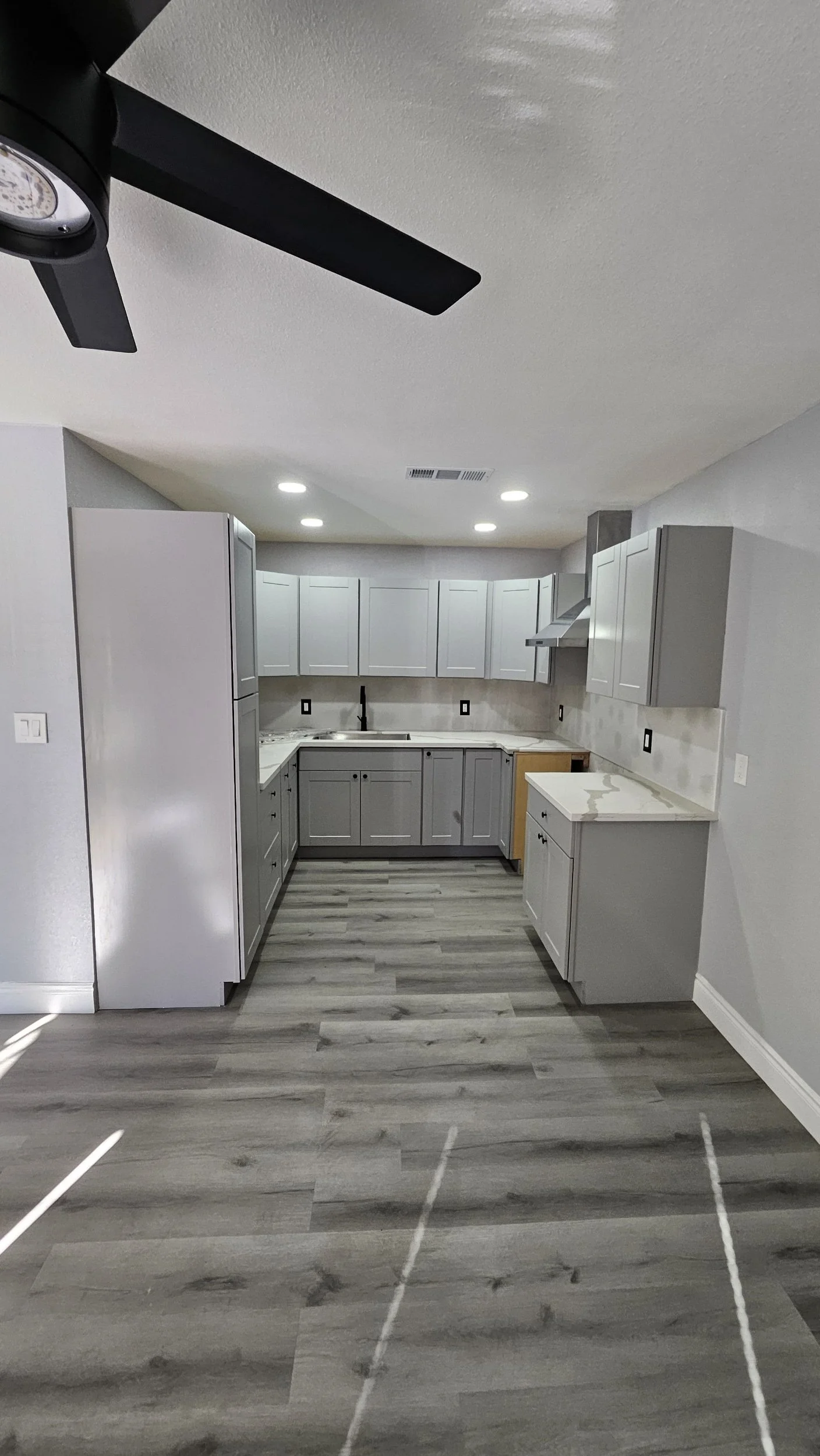 A modern kitchen with light gray cabinets, a marble countertop, gray patterned flooring, and ceiling lighting. There is a stove with a vent hood and a black faucet over the sink.