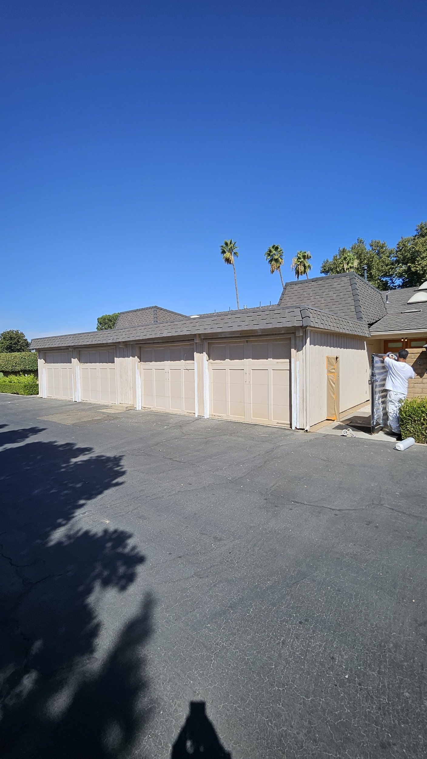 A person working on painting or staining a beige garage door on a residential property under a clear blue sky, with palm trees in the background.