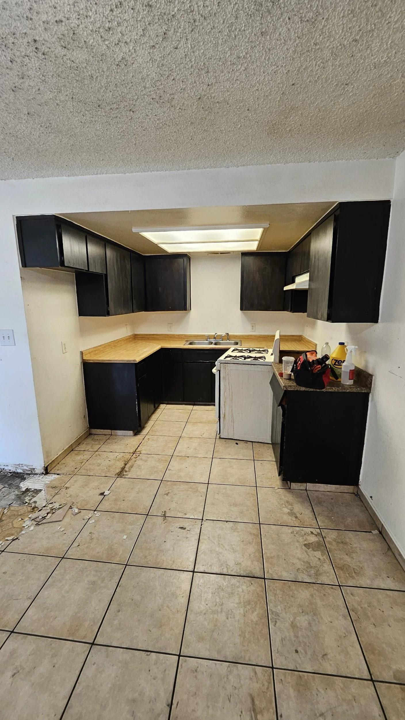 Renovated kitchen with black cabinets, beige countertops, a white stove, and a double sink with a window above. The floor has beige tiles, some dirt and debris, and cleaning supplies are on the counter.