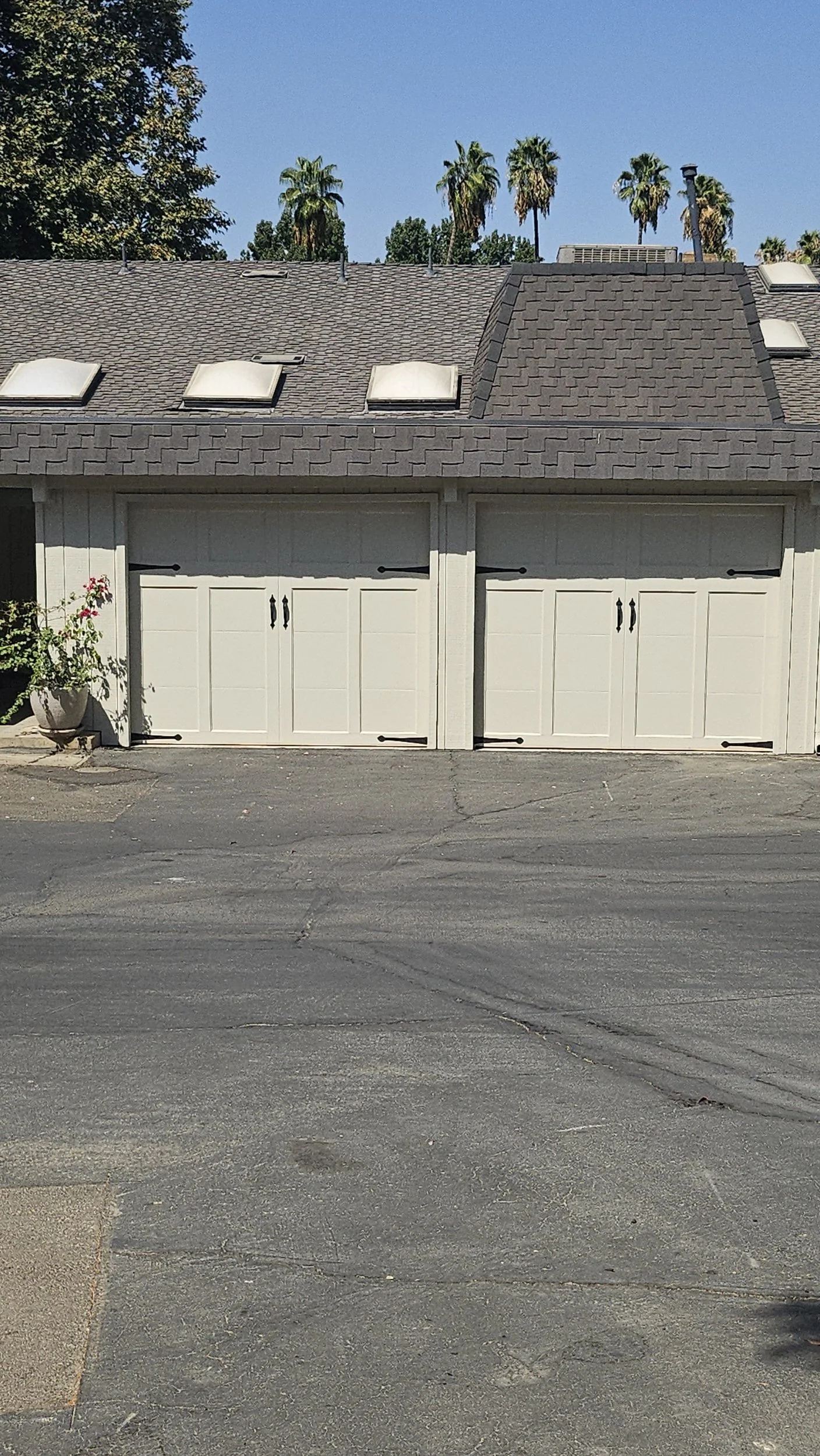 A two-car garage with white doors in a suburban neighborhood, with a roof featuring three skylights and four roof vents, and palm trees and other trees in the background under a clear blue sky.