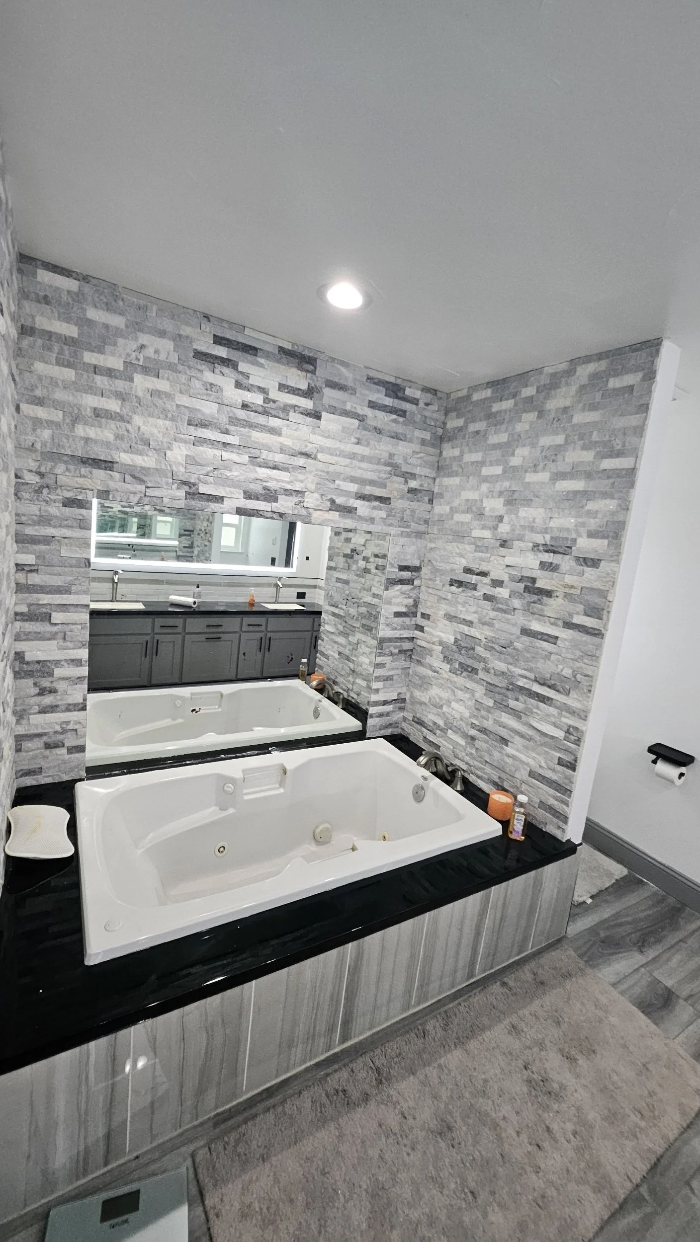 A bathroom with two white whirlpool tubs, grey stone tiled walls, a large mirror, dark grey cabinetry, and a window above the vanity area.