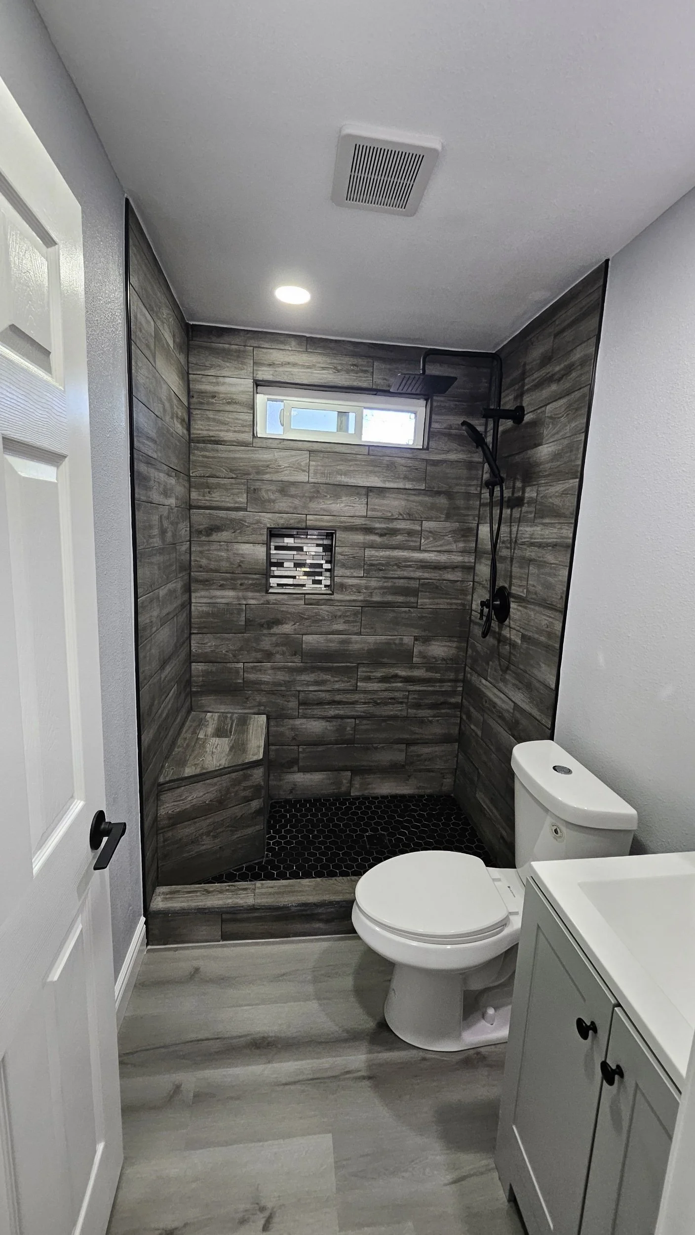 Bathroom with a walk-in shower featuring wood-look tile, a small window, a bench, and a black showerhead, along with a toilet and a gray vanity.