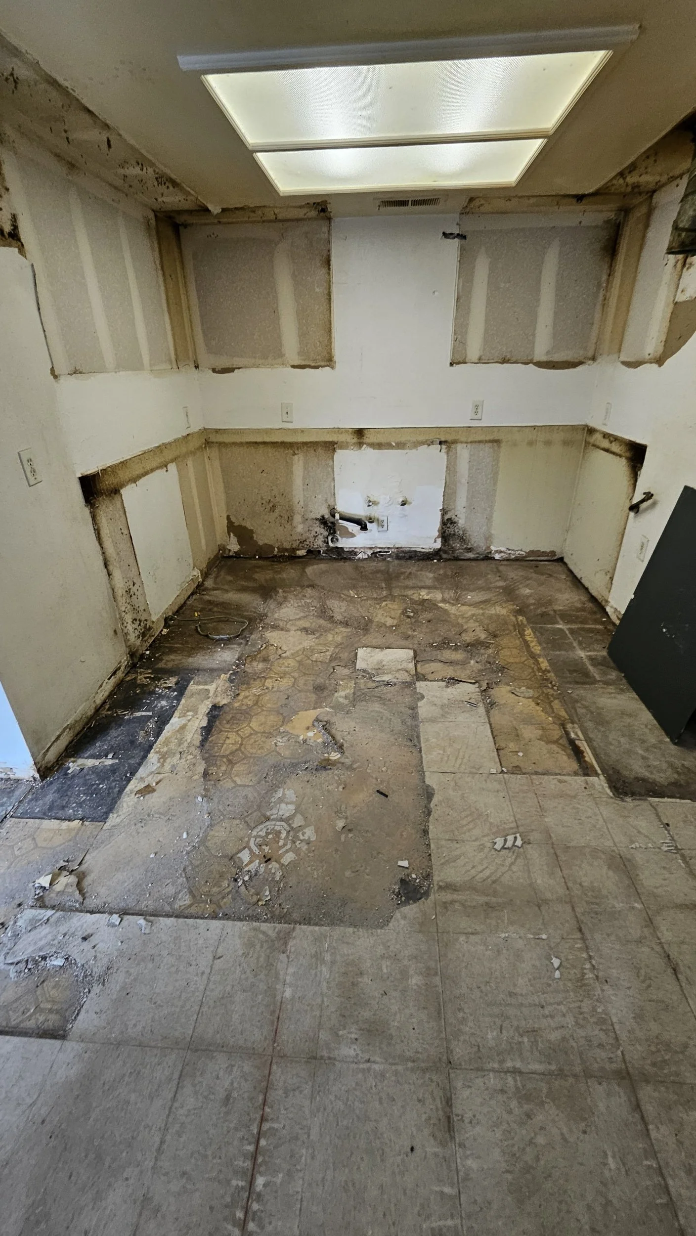 This is a kitchen or laundry room space in need of renovation, showing damaged walls, worn flooring, and exposed pipes.