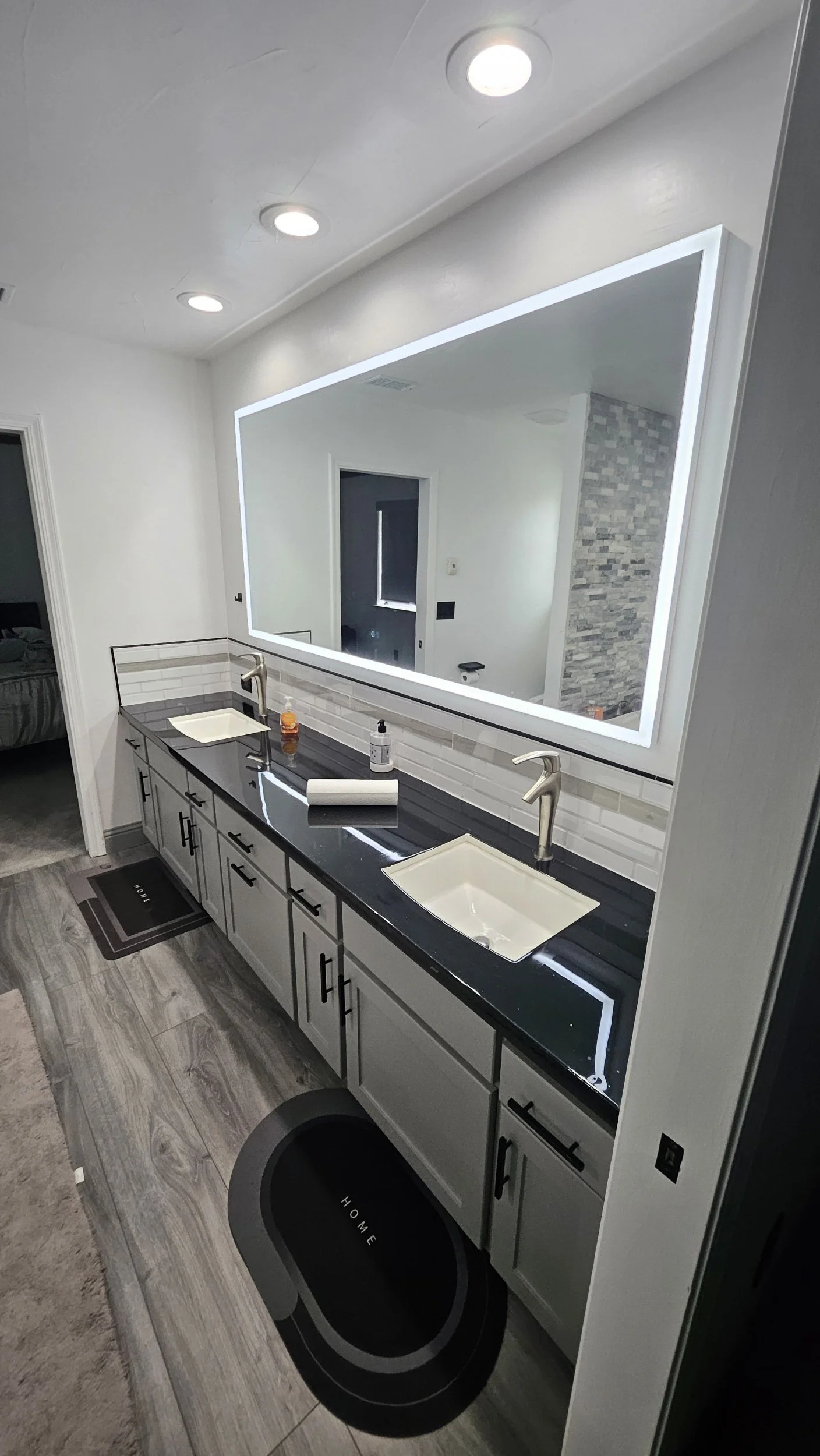 A modern bathroom vanity with a large illuminated mirror, double sinks, and a black countertop. The vanity has white cabinets with black handles. On the countertop, there are soap dispensers, hand sanitizer, and paper towels. A black mat is in front of the vanity, and a pet food bowl labeled 'HOME' is on the floor. Recessed ceiling lights and a gray stone accent wall are visible in the reflection of the mirror.