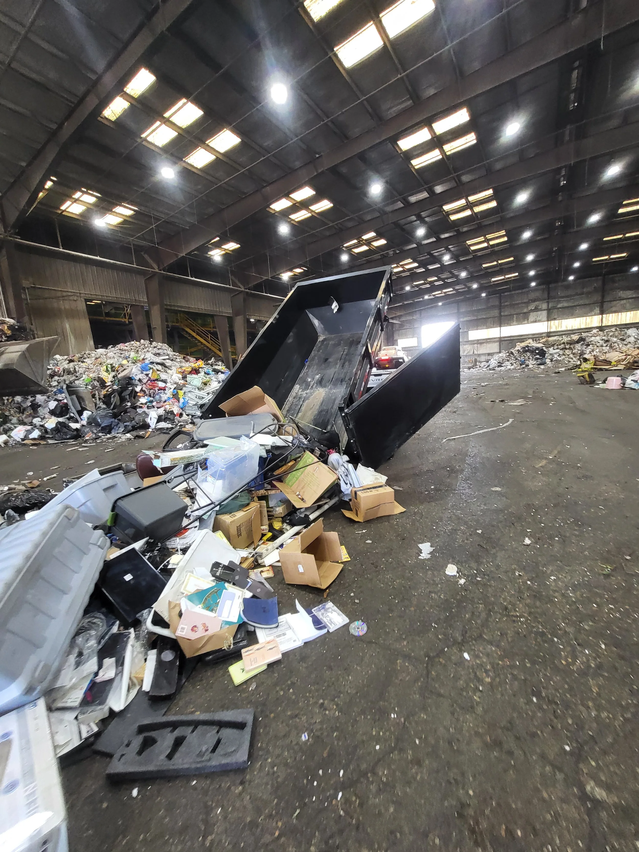 Interior of a warehouse or recycling center with piles of mixed trash and cardboard, and a large black dumpster tipped over.