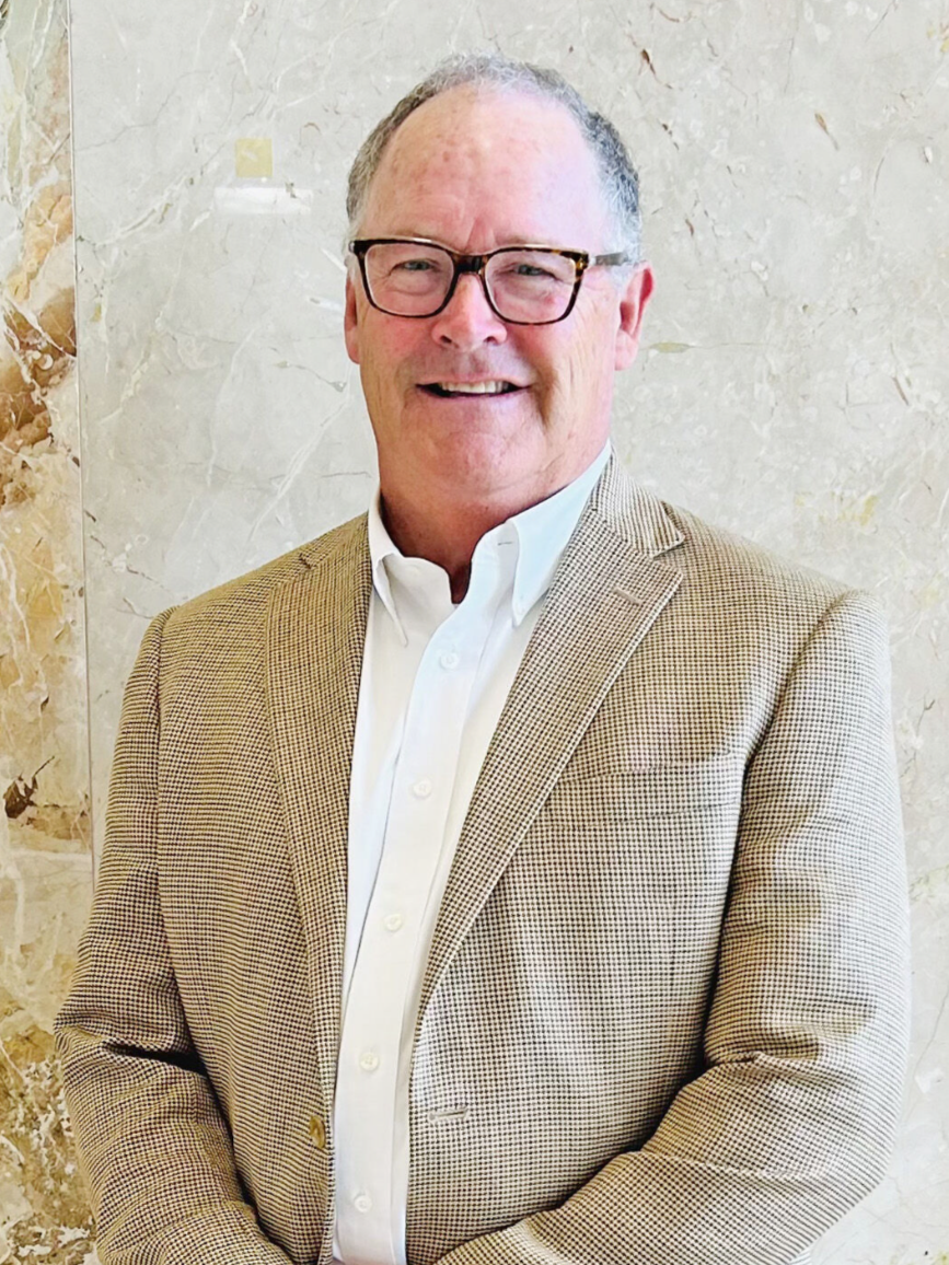 A middle-aged man wearing glasses, a white dress shirt, and a beige checkered blazer, smiling and standing in front of a beige marble wall.