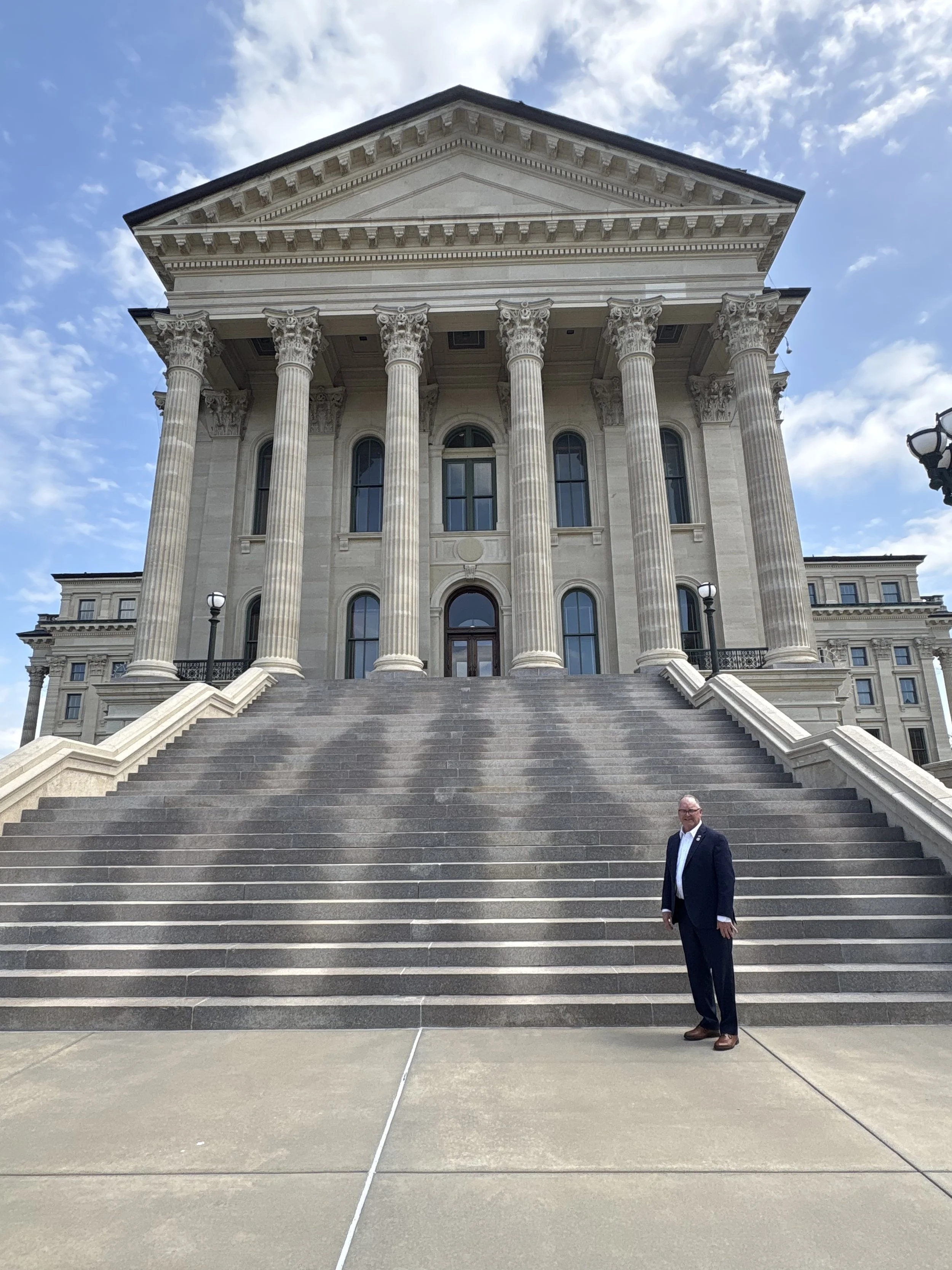 A man in a suit standing at the bottom of large stairs in front of a neoclassical government building with tall columns and an ornate facade, under a partly cloudy sky.