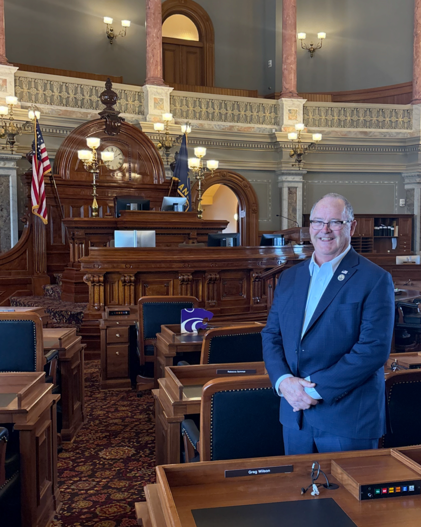 A man in a suit and glasses standing in a room with ornate woodwork, chandeliers, American flags, and leather chairs.